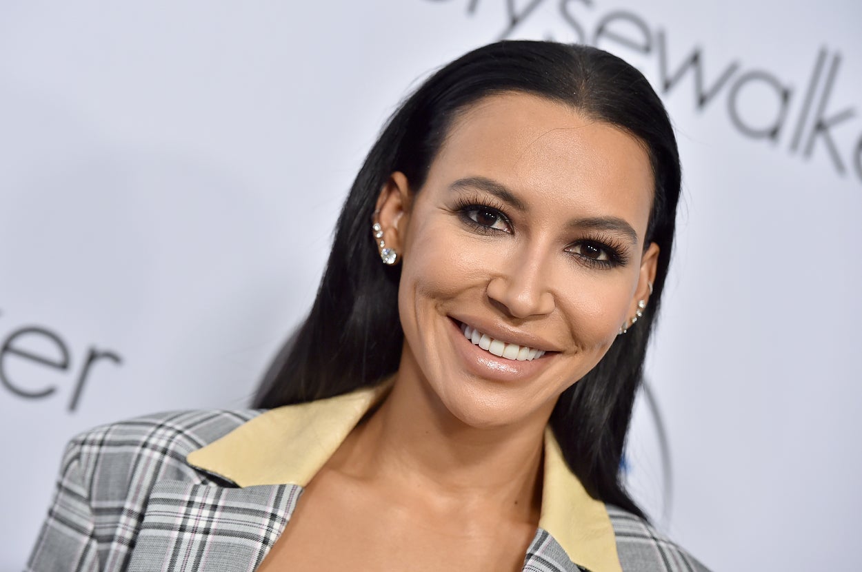 Naya Rivera smiling at an event, wearing a checkered blazer with a yellow collar and earring on one ear
