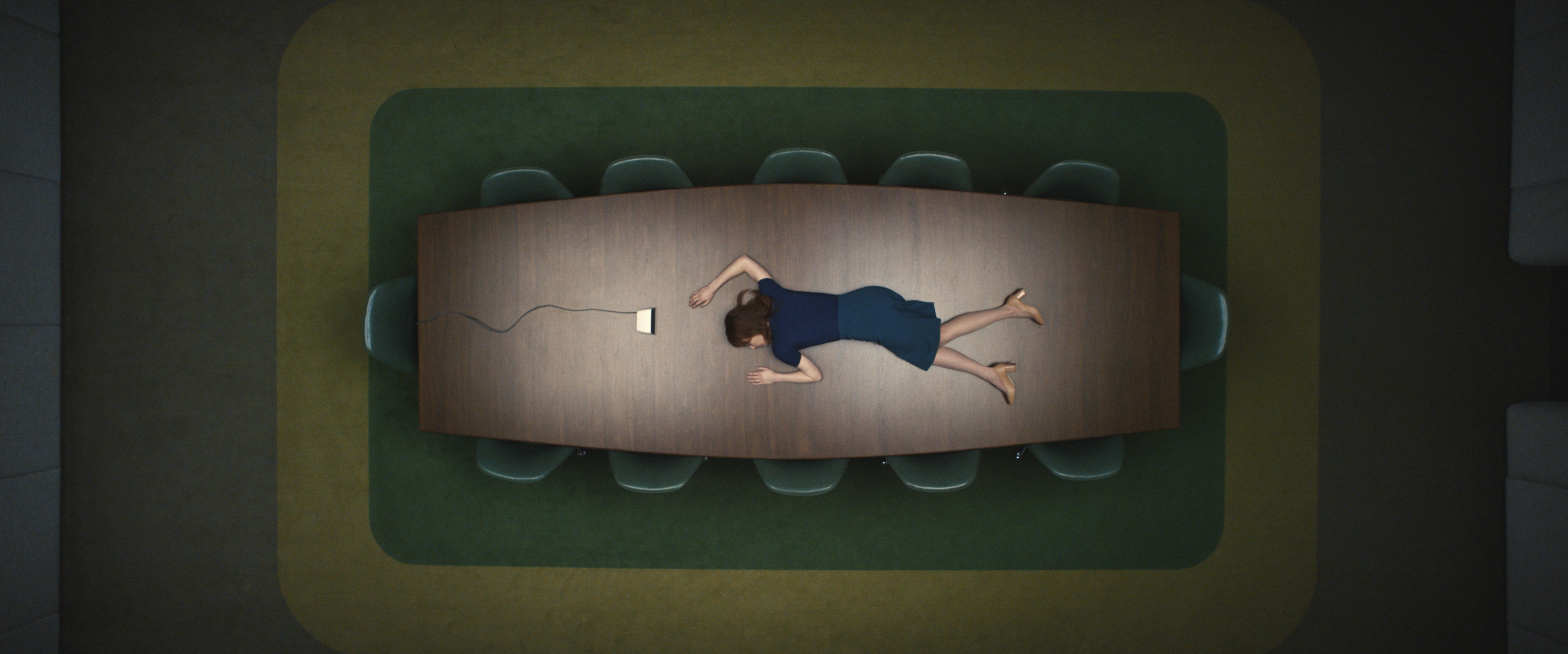Person in business attire lies face down on a conference table, viewed from above, with a styrofoam cup nearby