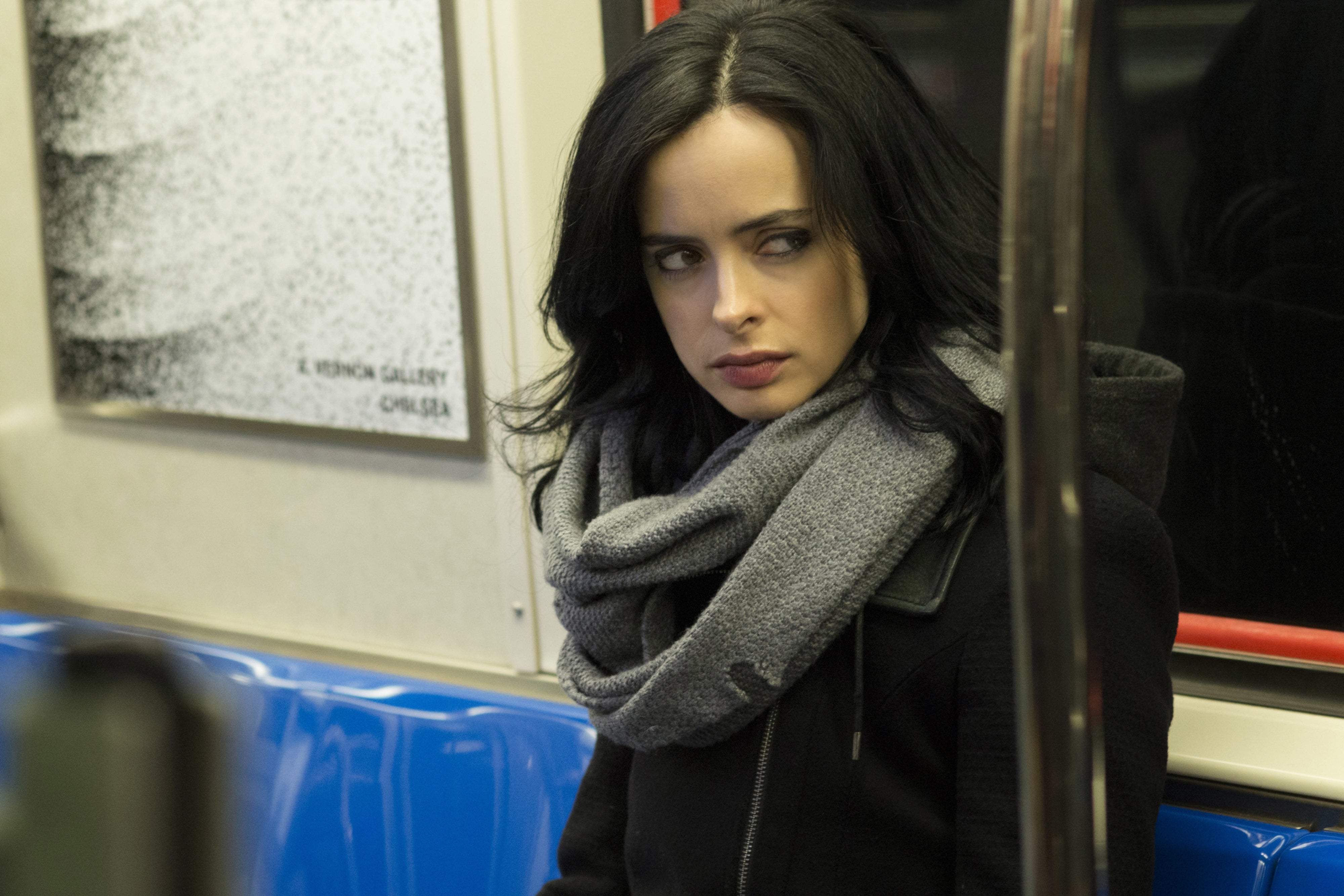 Woman on a subway with long dark hair, wearing a gray scarf and black coat, looking to the side with a thoughtful expression