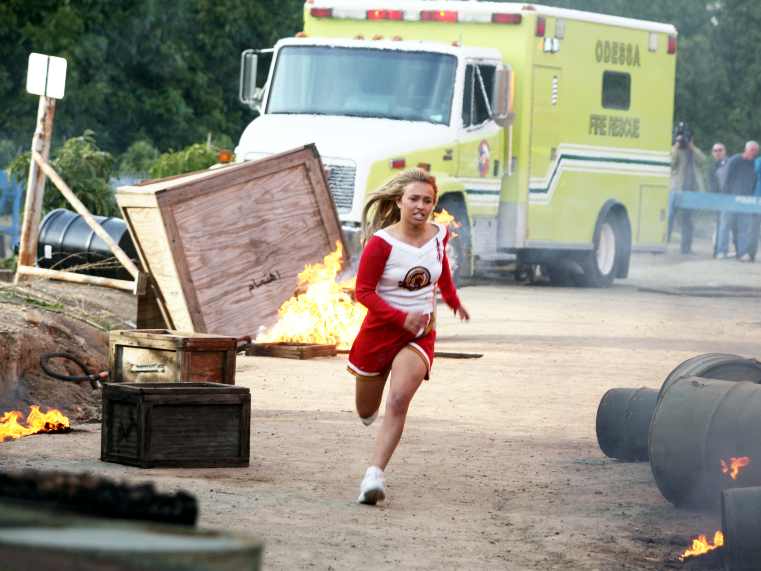 Person in cheerleader outfit runs through an obstacle course with fire and debris, while an emergency vehicle is seen in the background