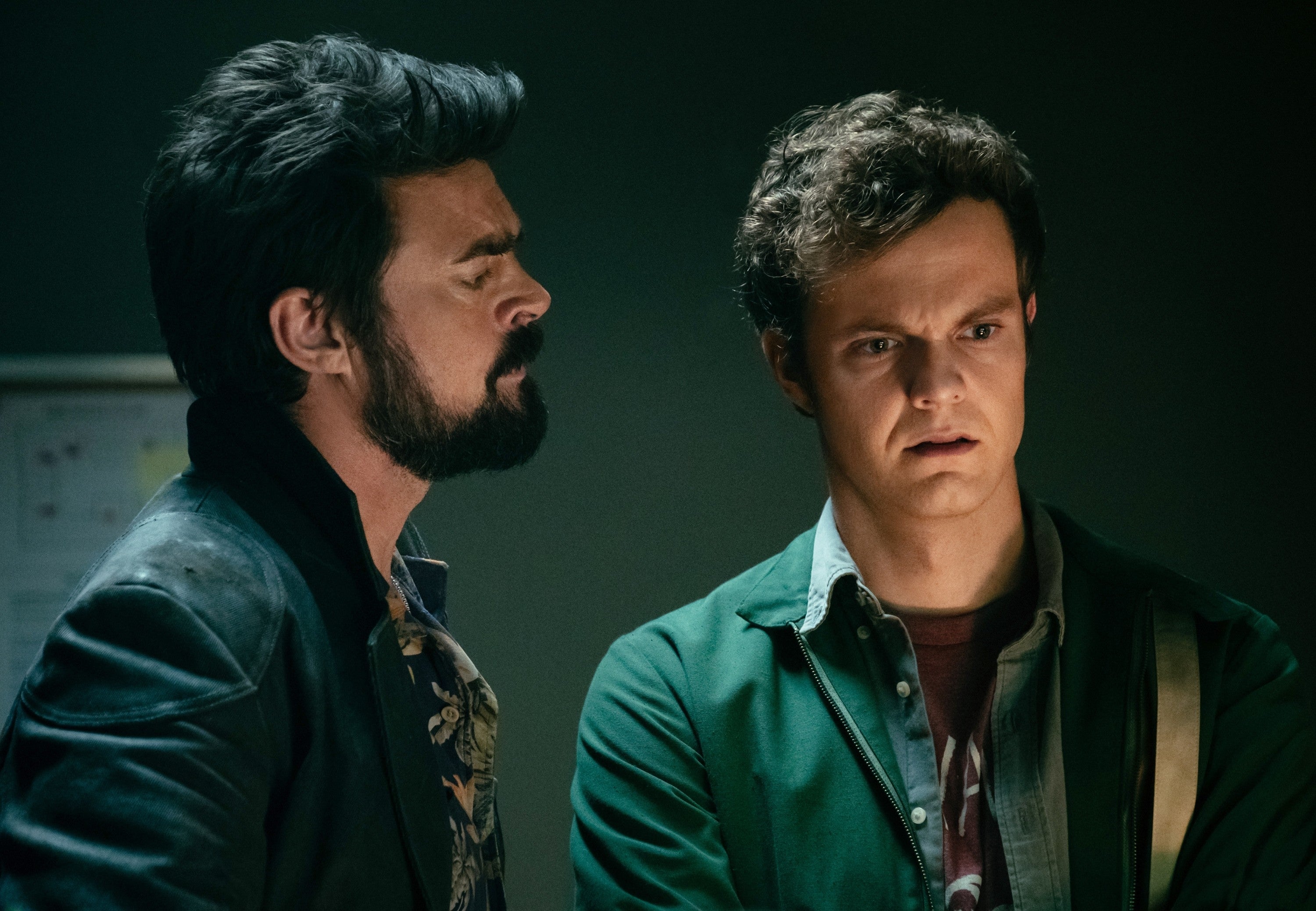 Two men stand close; one in a floral shirt under a jacket, the other in a casual shirt and jacket, appearing serious, possibly discussing something