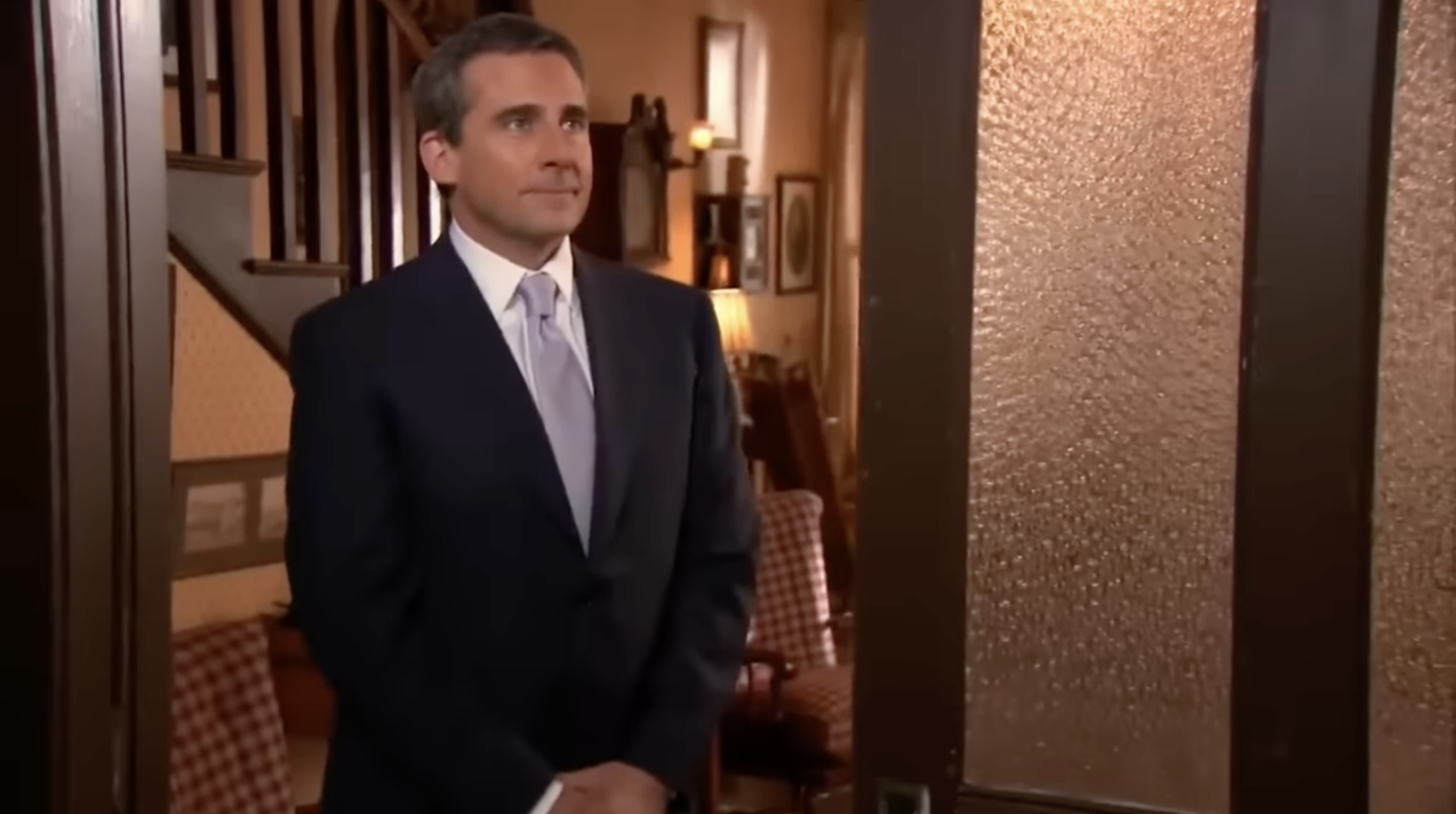 A man in a suit stands indoors, looking contemplative, as if entering a room. Background shows chairs and a staircase