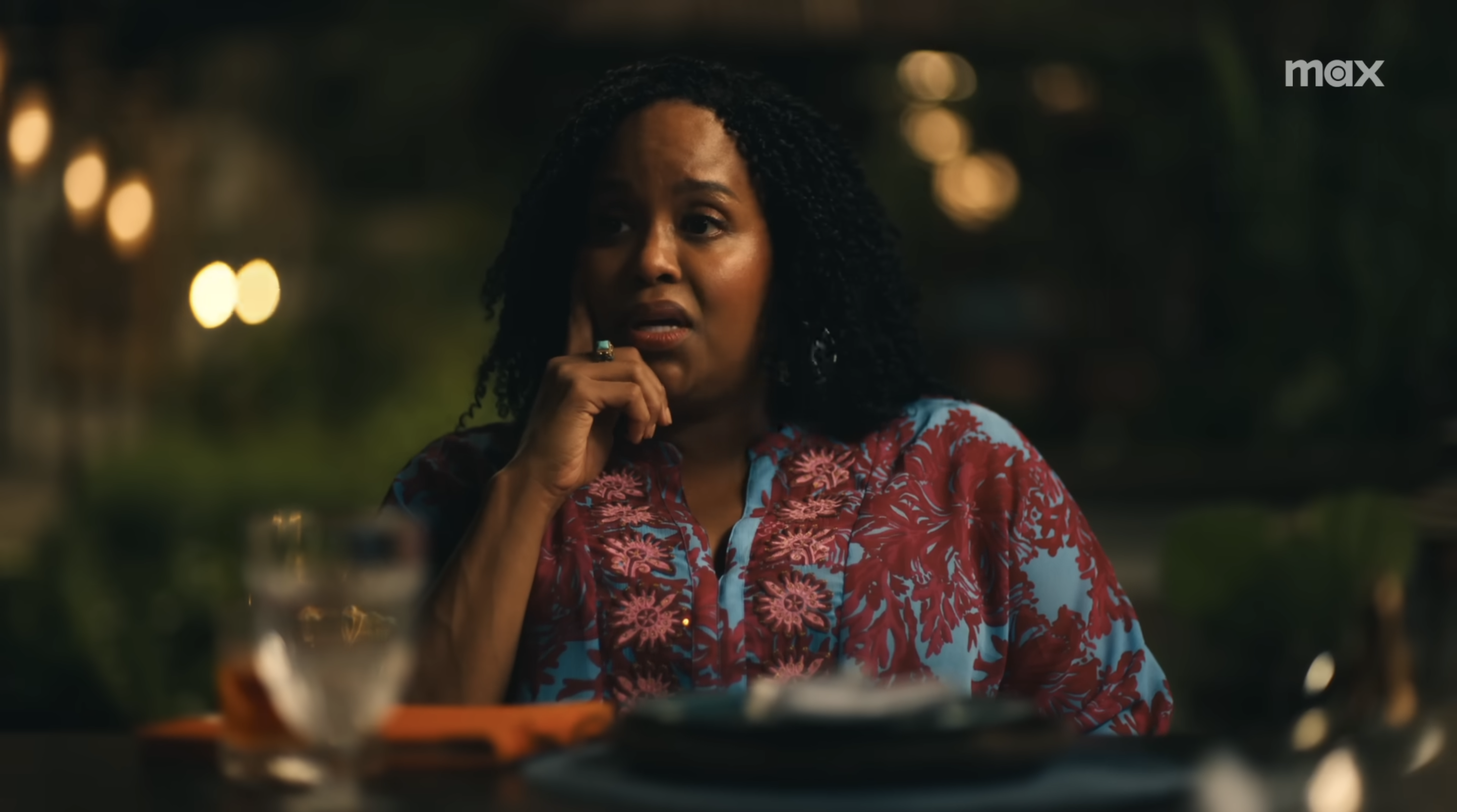 A person seated at a dining table, wearing a floral-patterned shirt, appears to be engaged in conversation