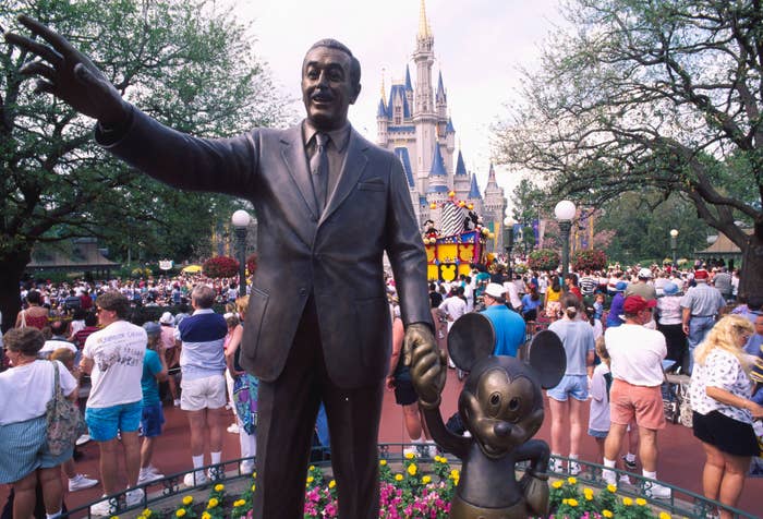 Statue of a famous man with an iconic mouse character in front of a castle, surrounded by a large crowd at a theme park