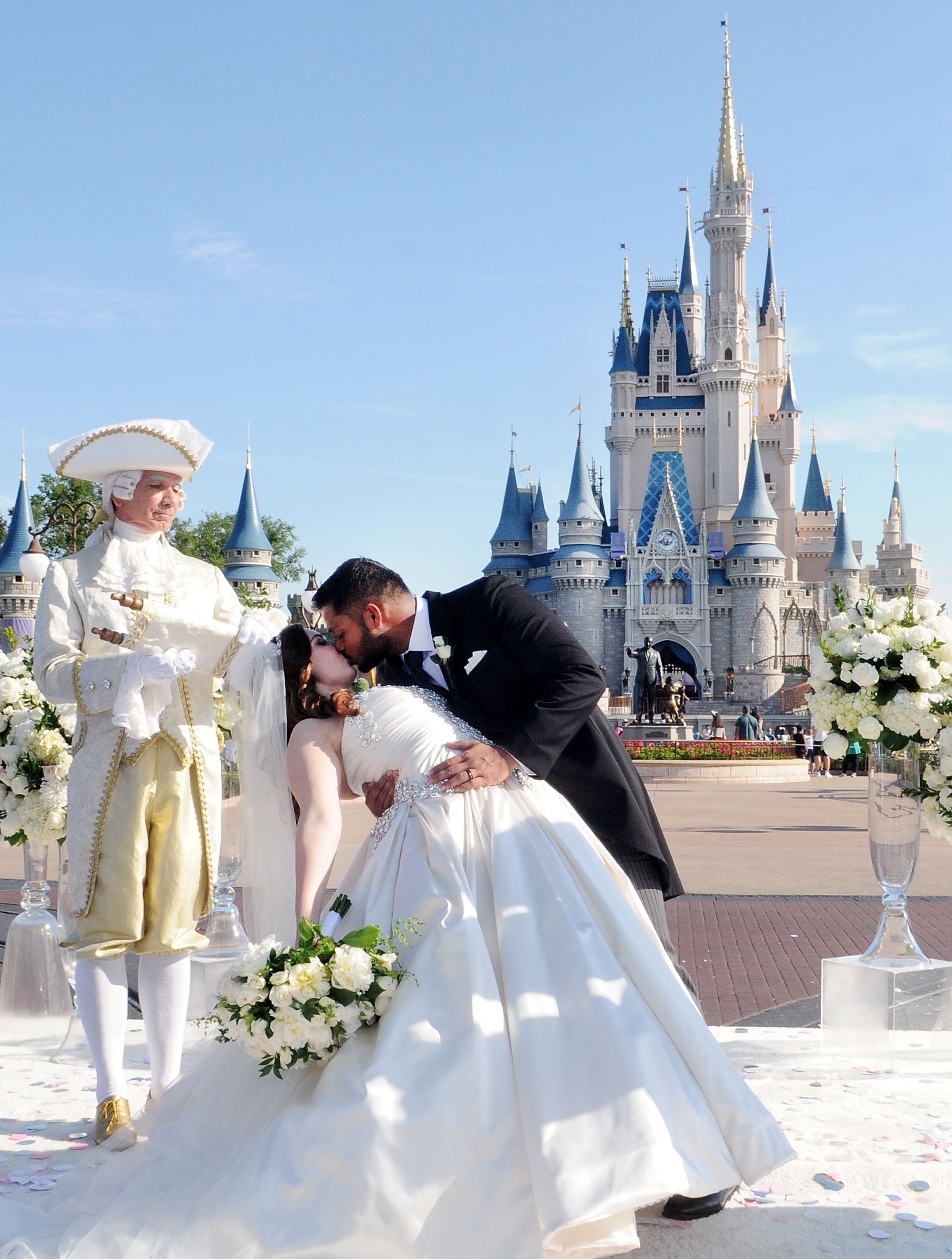 Couple in formal attire kiss in front of a castle, observed by a man in historical costume, set against a theme park backdrop