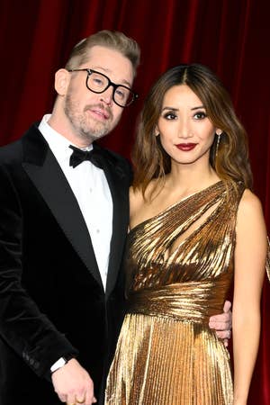 Two people on the red carpet; one in a black tuxedo and bow tie, and the other in a shimmering gold gown with an elegant drape