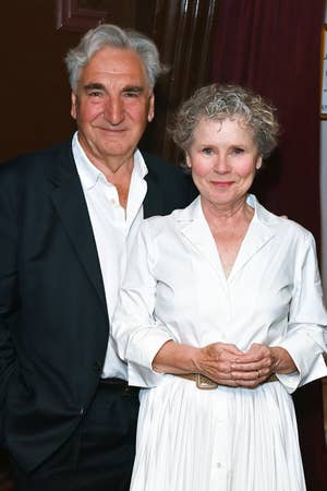 Two people in formal wear stand together, smiling. One is in a suit, and the other wears a long, elegant white dress