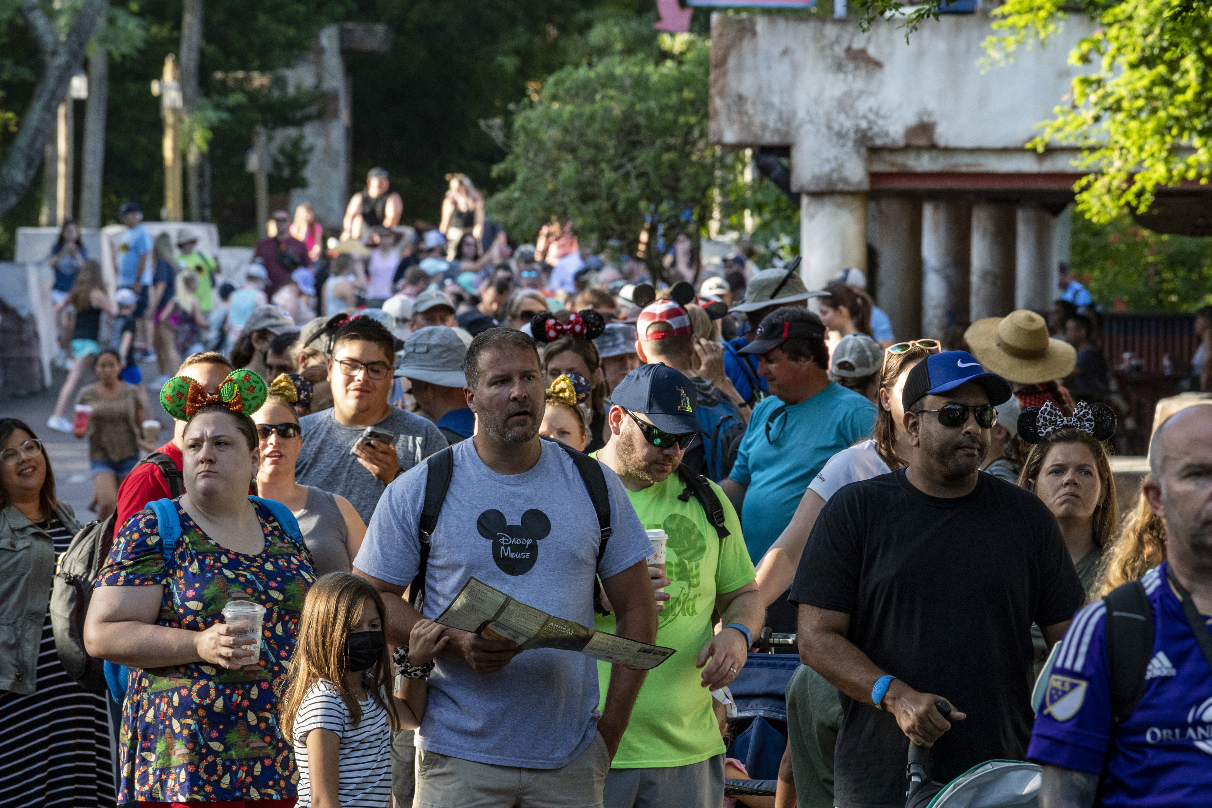 A large, diverse crowd lines an outdoor theme park pathway, some wearing Disney-themed attire and hats. They appear to be waiting or moving forward