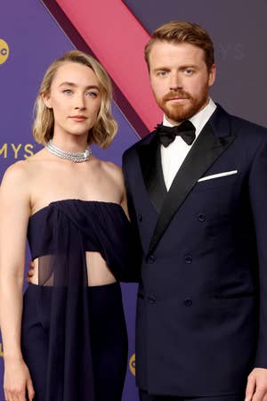 Two individuals on a red carpet. The woman wears a strapless dress with a sheer panel and choker necklace. The man is in a tailored double-breasted tuxedo