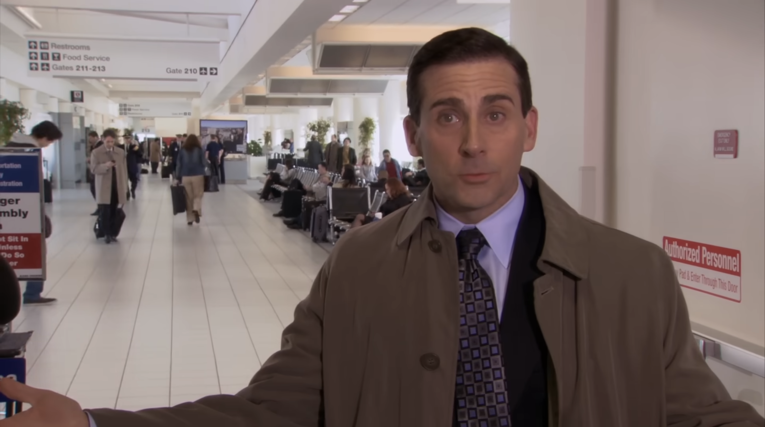 Person in a busy airport hallway, pointing, wearing a trench coat and suit; people are seated or walking in the background