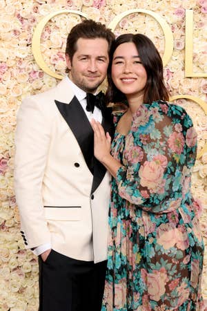 A man in a white tuxedo and a woman in a floral sheer dress pose together in front of a floral backdrop at a formal event