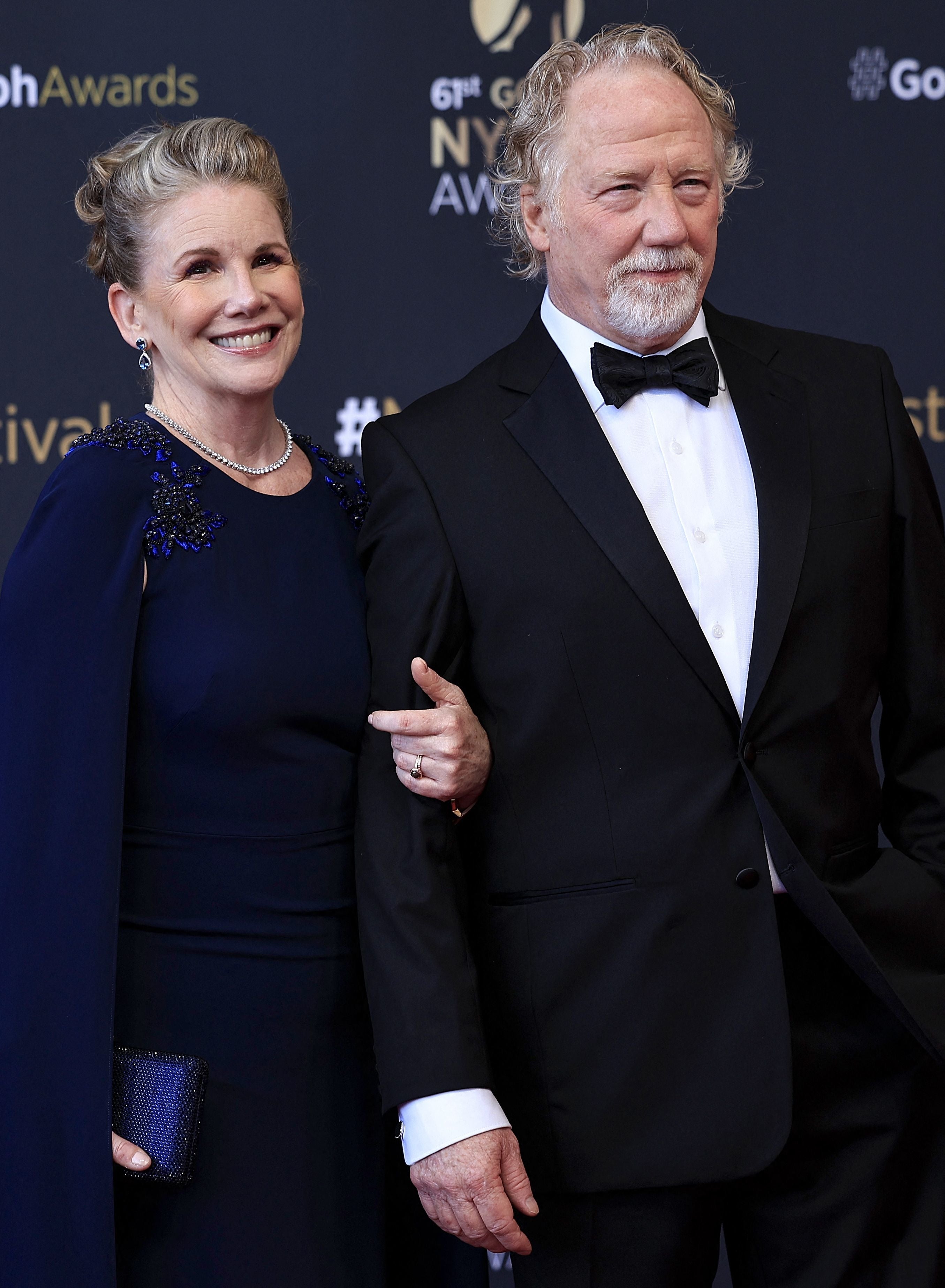 A smiling couple poses on the red carpet. The woman wears an elegant gown with embellishments, and the man is in a classic black tuxedo