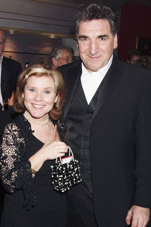 Two people in formal black attire smiling at an event, surrounded by others. One holds a beaded clutch and a drink