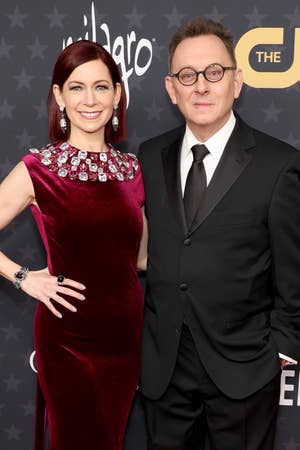 Two people posing on a red carpet; the woman is wearing a velvet dress with jeweled neckline, and the man is in a classic black suit and tie