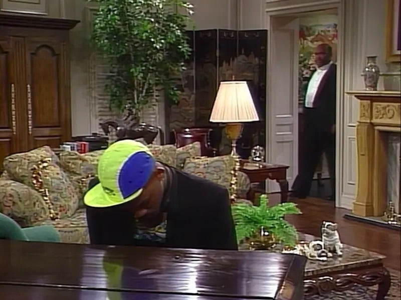 Person sits at a grand piano in a luxurious living room, wearing a cap with a flipped brim. Another person in formal wear stands in the doorway