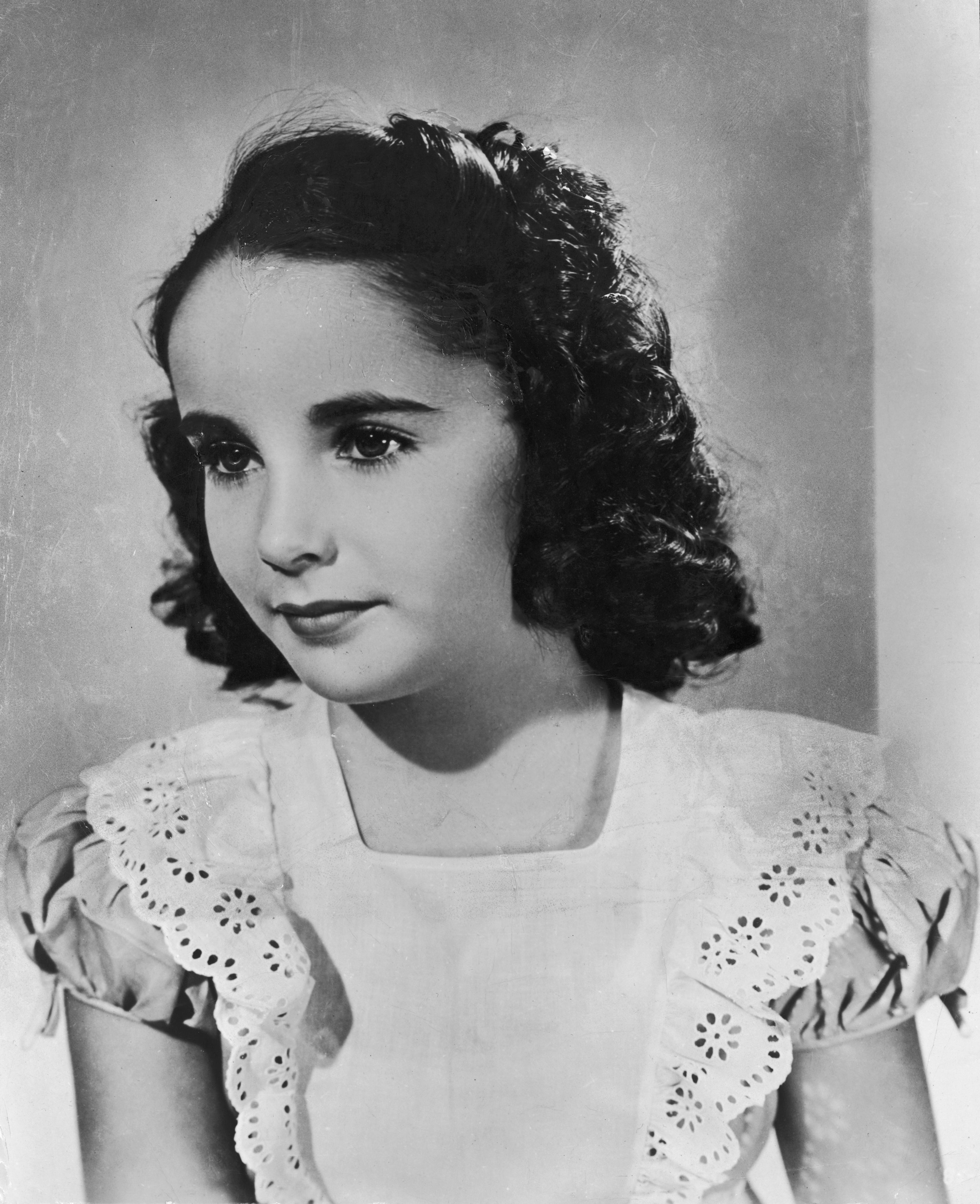A young girl with dark curly hair wears a vintage-style dress with lace details, looking contemplative