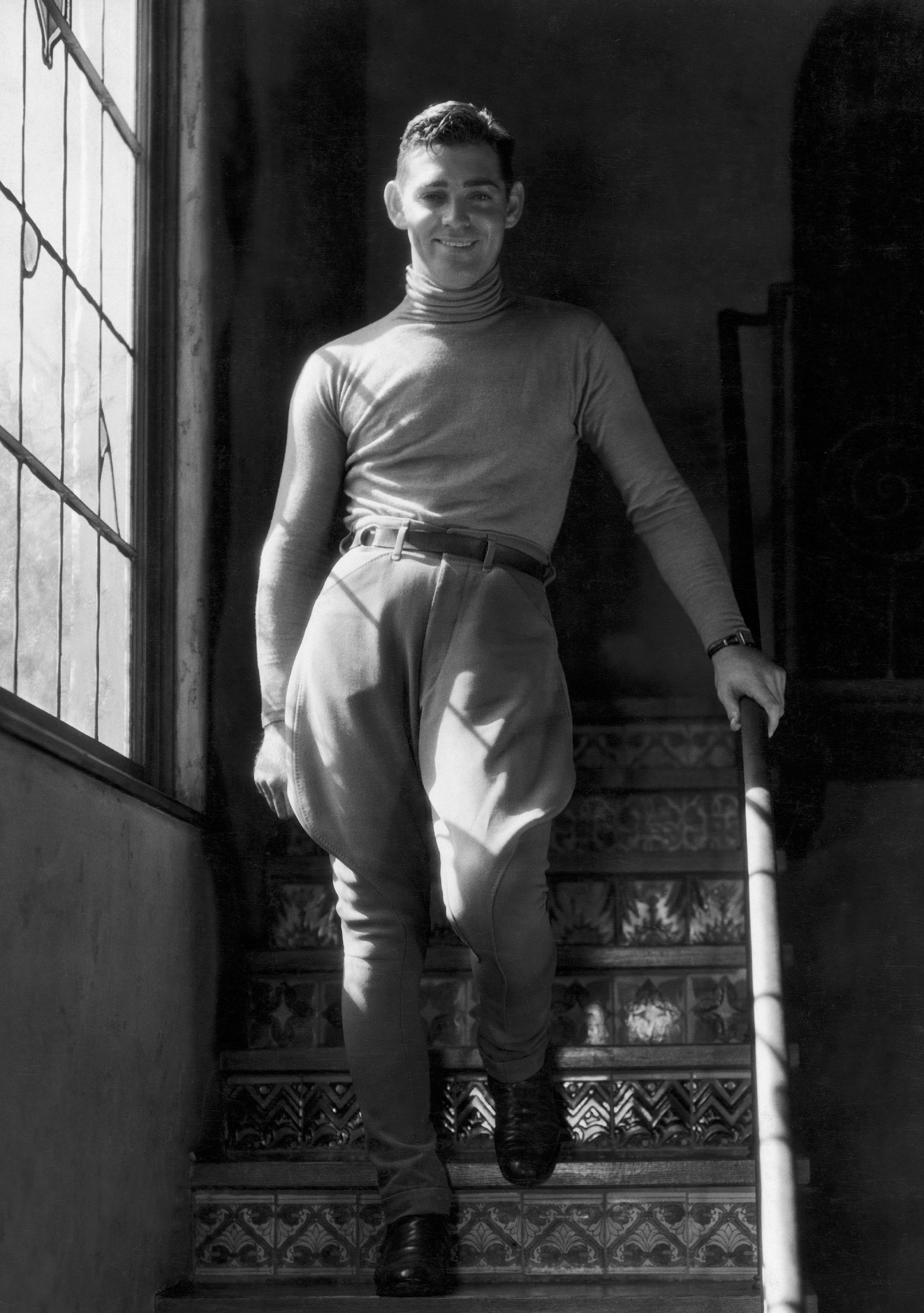 Person in vintage attire, wearing high-waisted pants and a turtleneck, stands on a decorative staircase, smiling