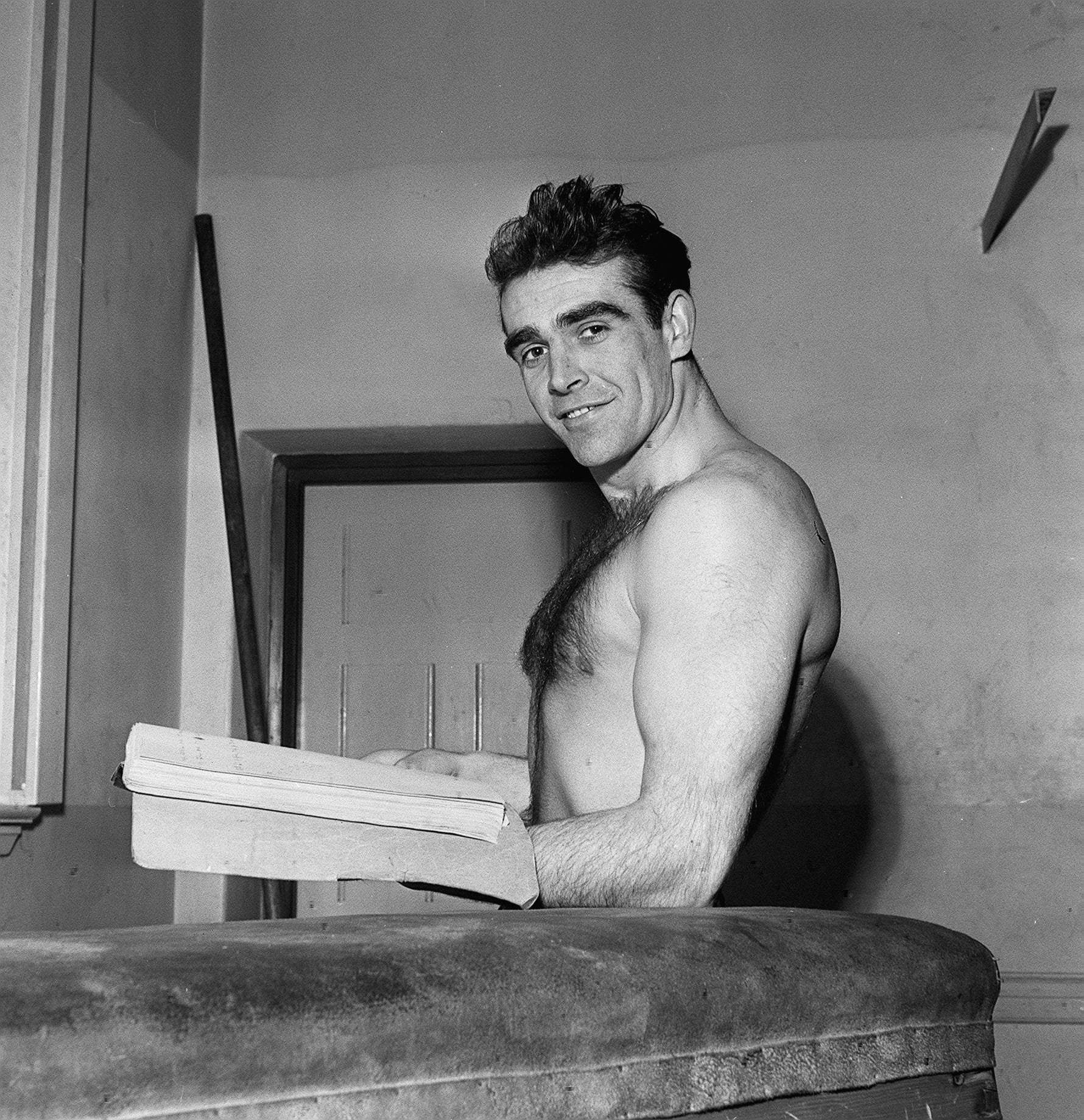 Man posing shirtless, holding open a book, standing indoors