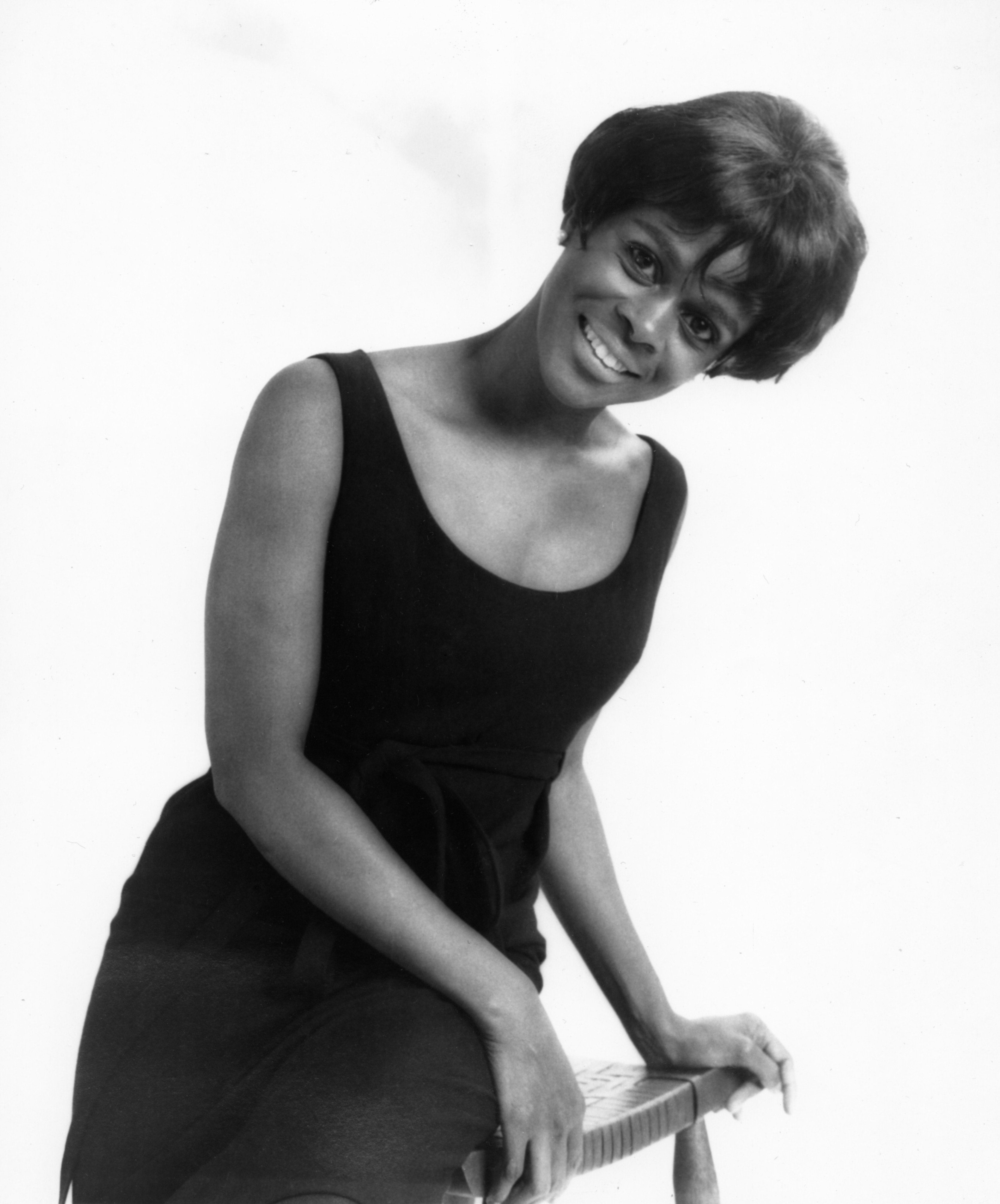 Person smiling in a classic black dress, seated on a chair, in a studio setting. Hair is styled short and elegant