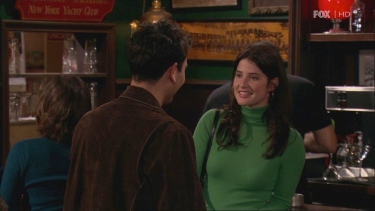 Two people chat in a bar; one woman wears a green turtleneck. The setting resembles a casual TV show scene