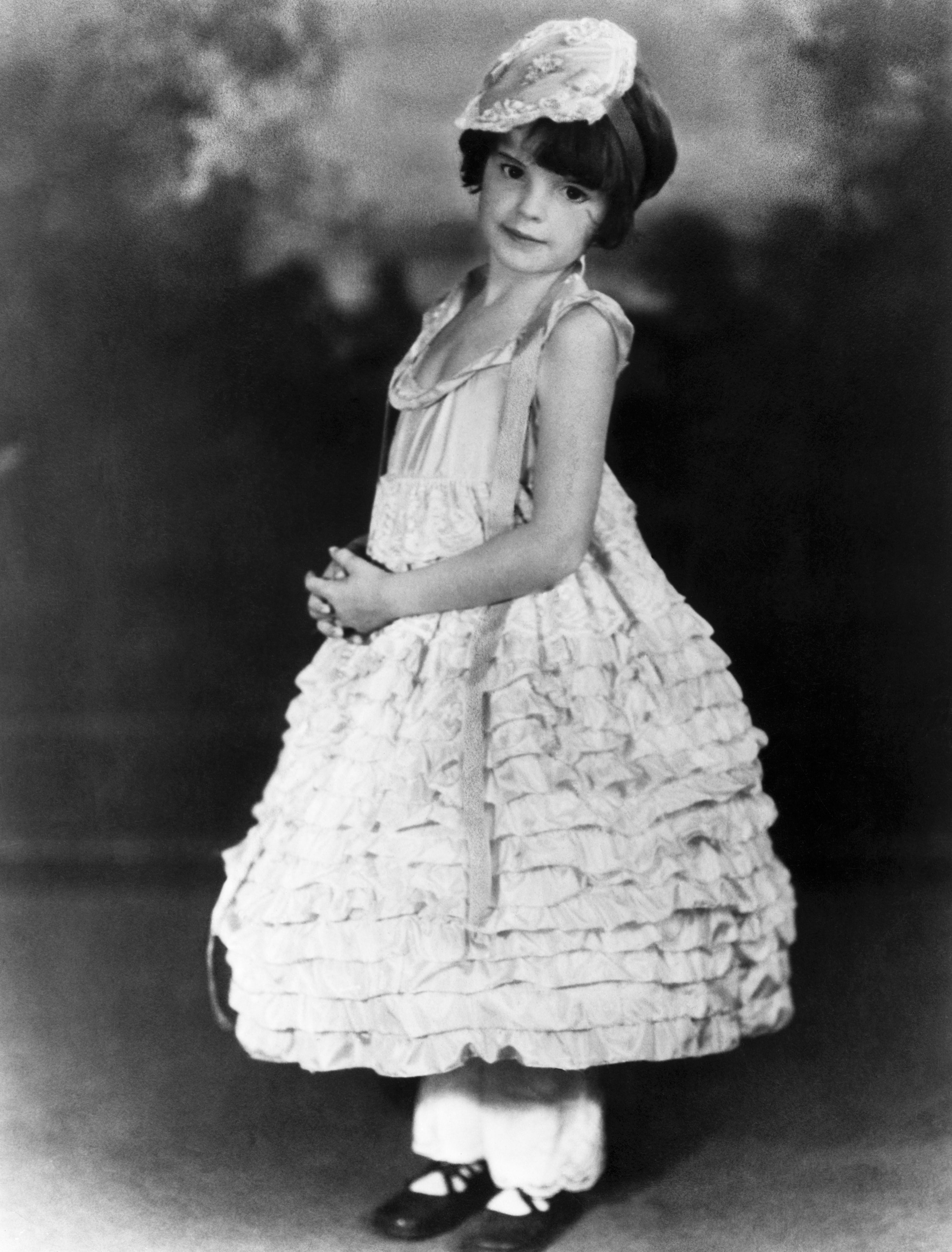 A young child in a frilly, ruffled dress and hat poses elegantly, evoking early 20th-century fashion