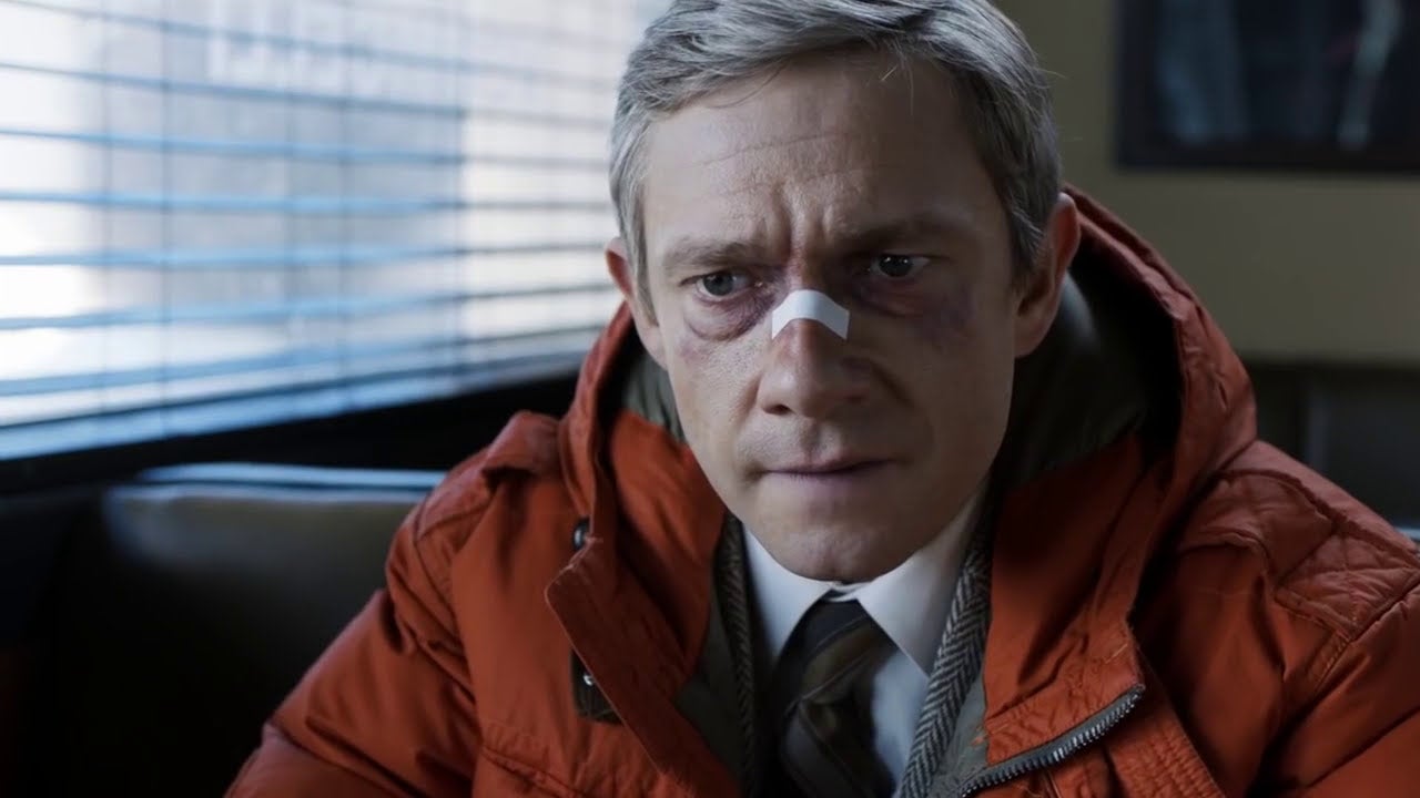 A serious-looking man with a nose bandage sits in a diner, wearing a hooded jacket and tie