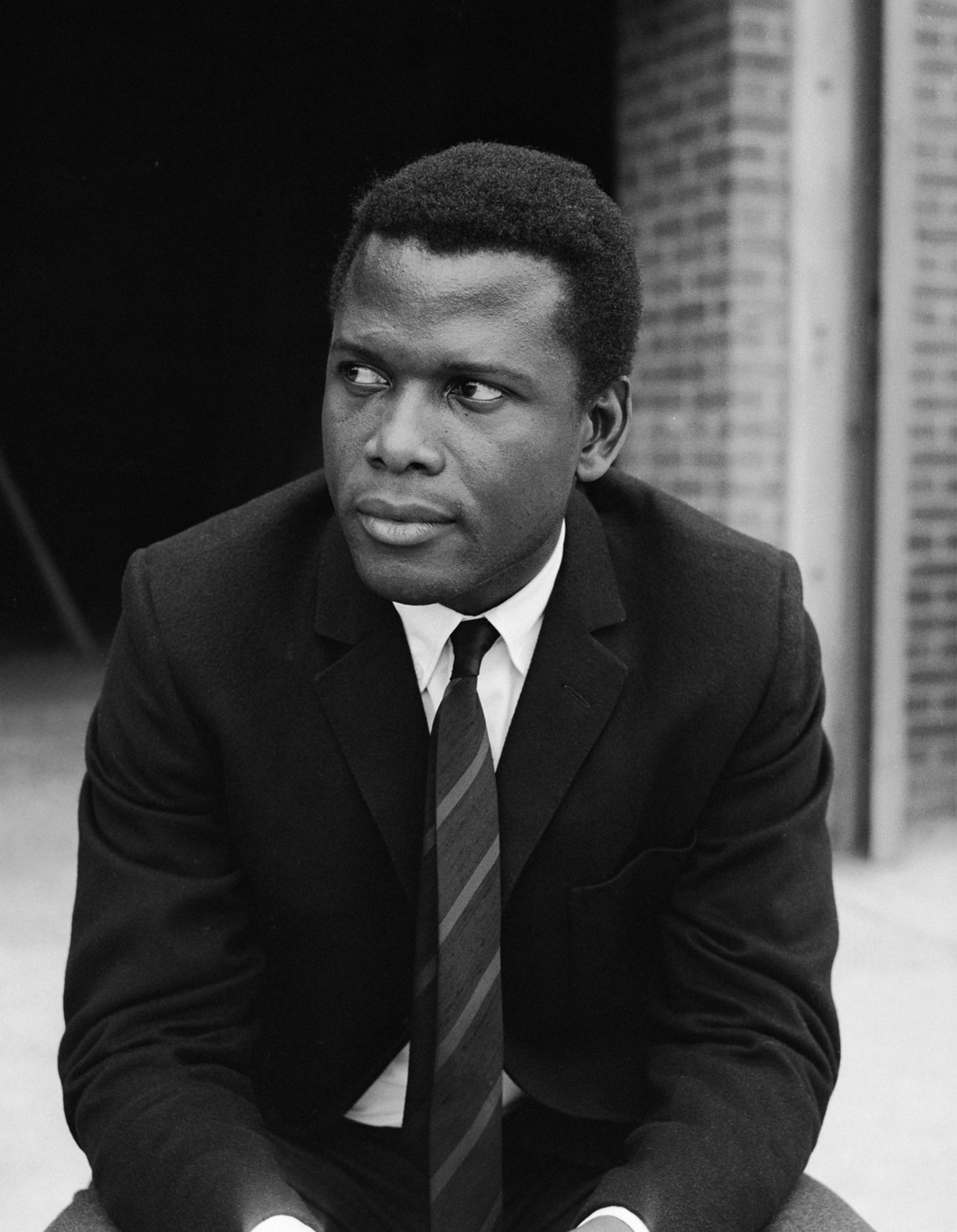 A person in a classic suit and tie sits thoughtfully, looking into the distance. The setting is a brick building
