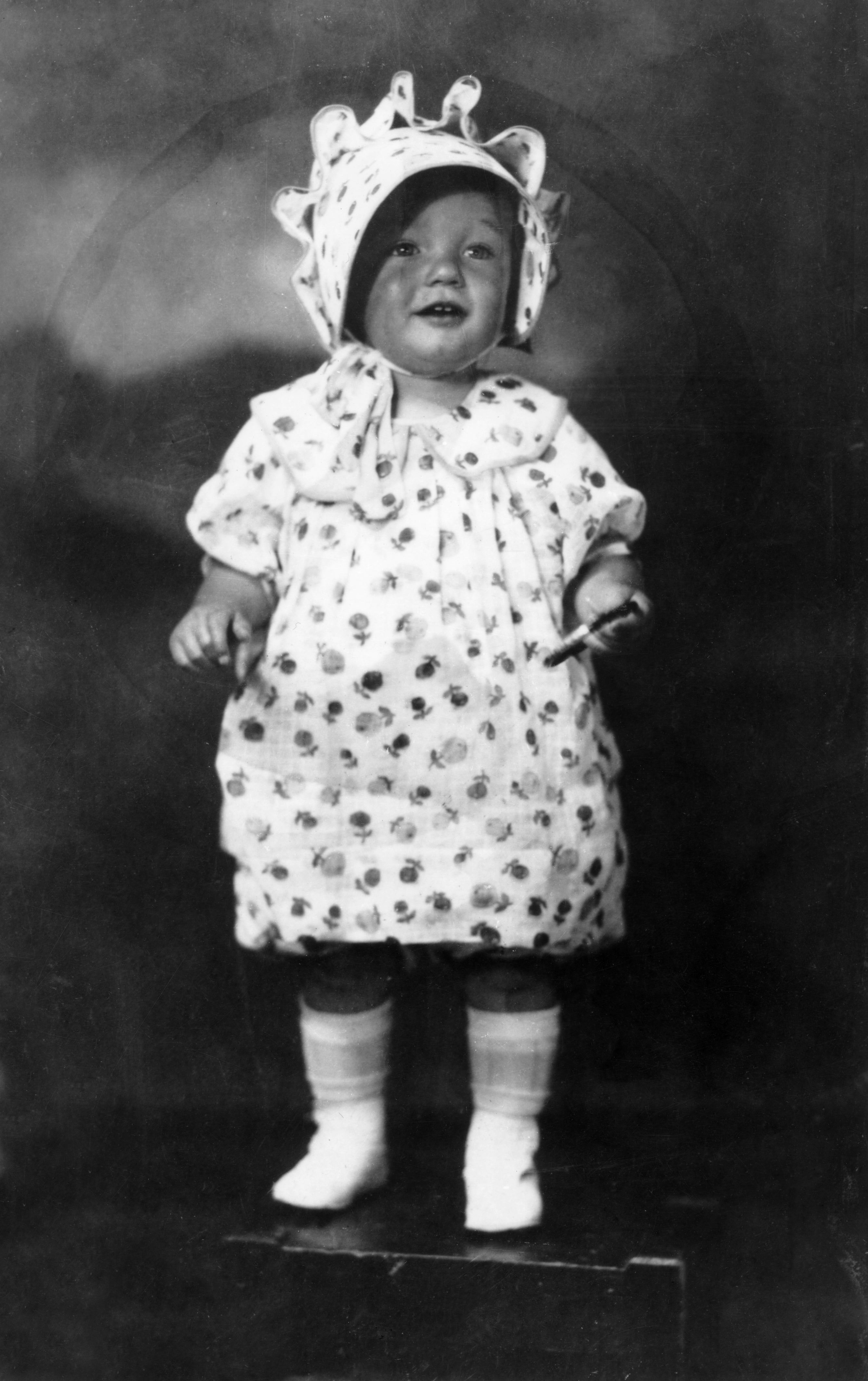 A vintage photo of a smiling baby in a patterned dress and bonnet, standing on a platform