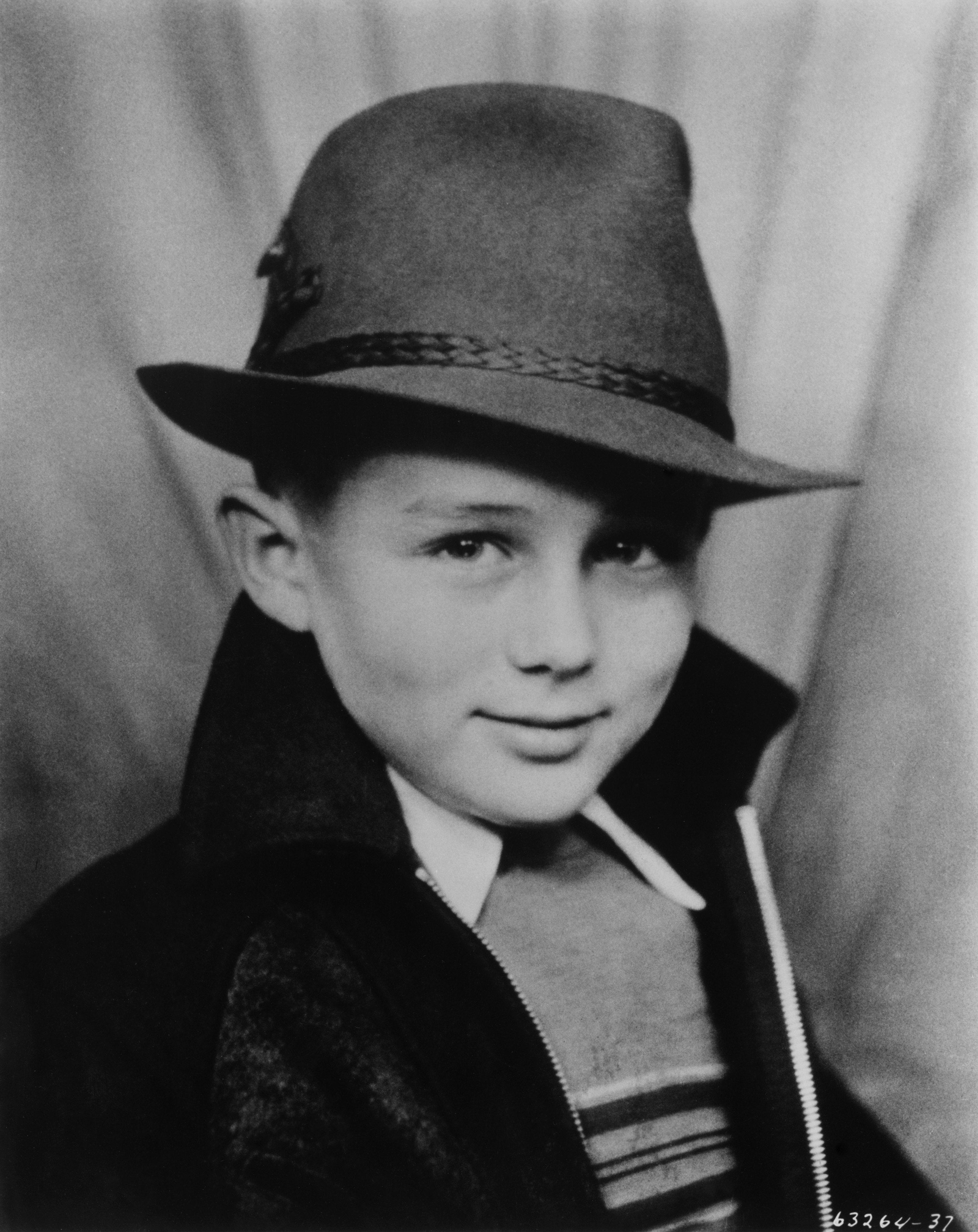 A young boy wearing a suit, shirt, and fedora, poses confidently for a portrait against a plain backdrop