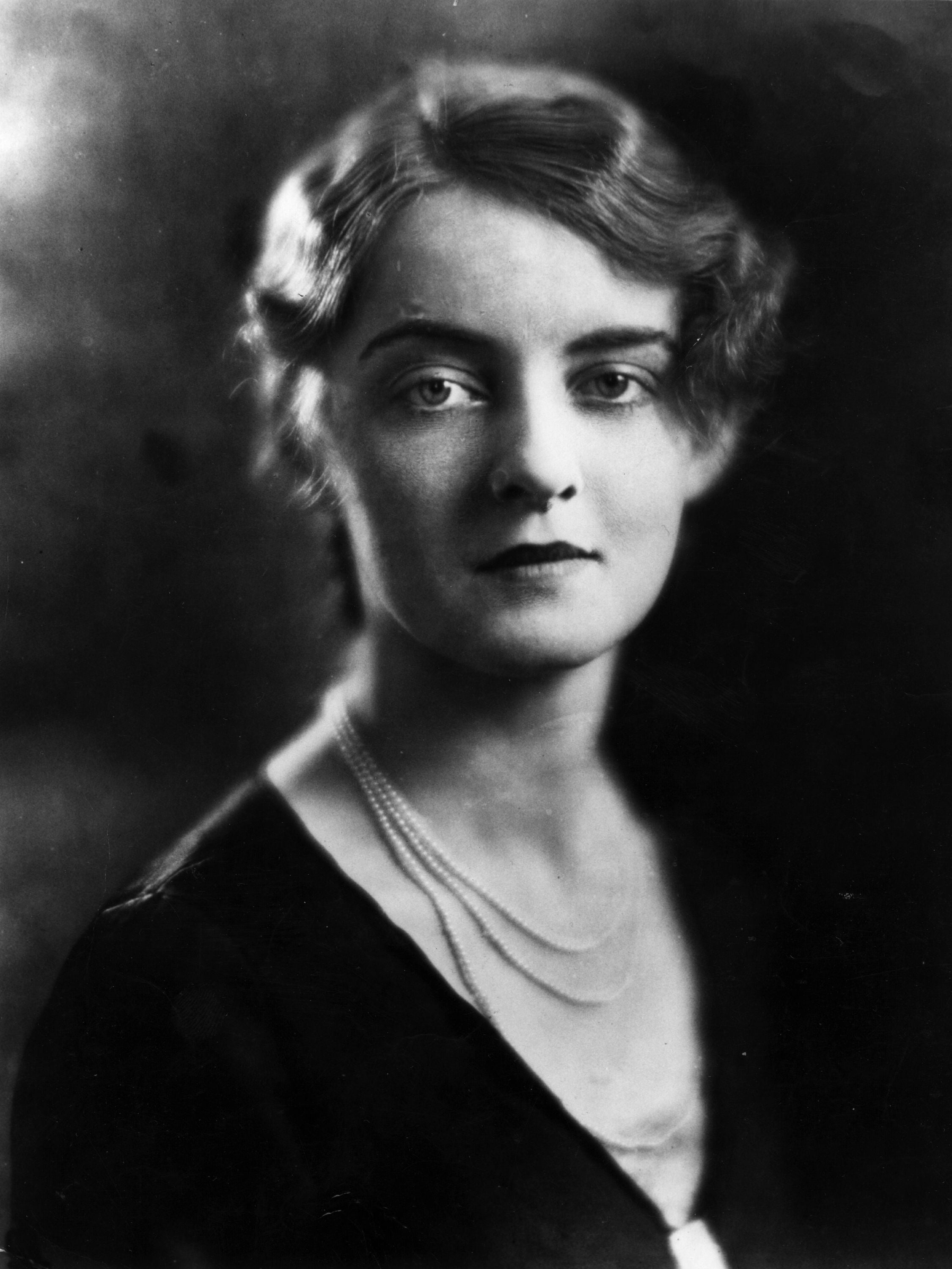 Person in vintage-style portrait with short wavy hair, wearing a necklace and a dark top, looks directly at the camera