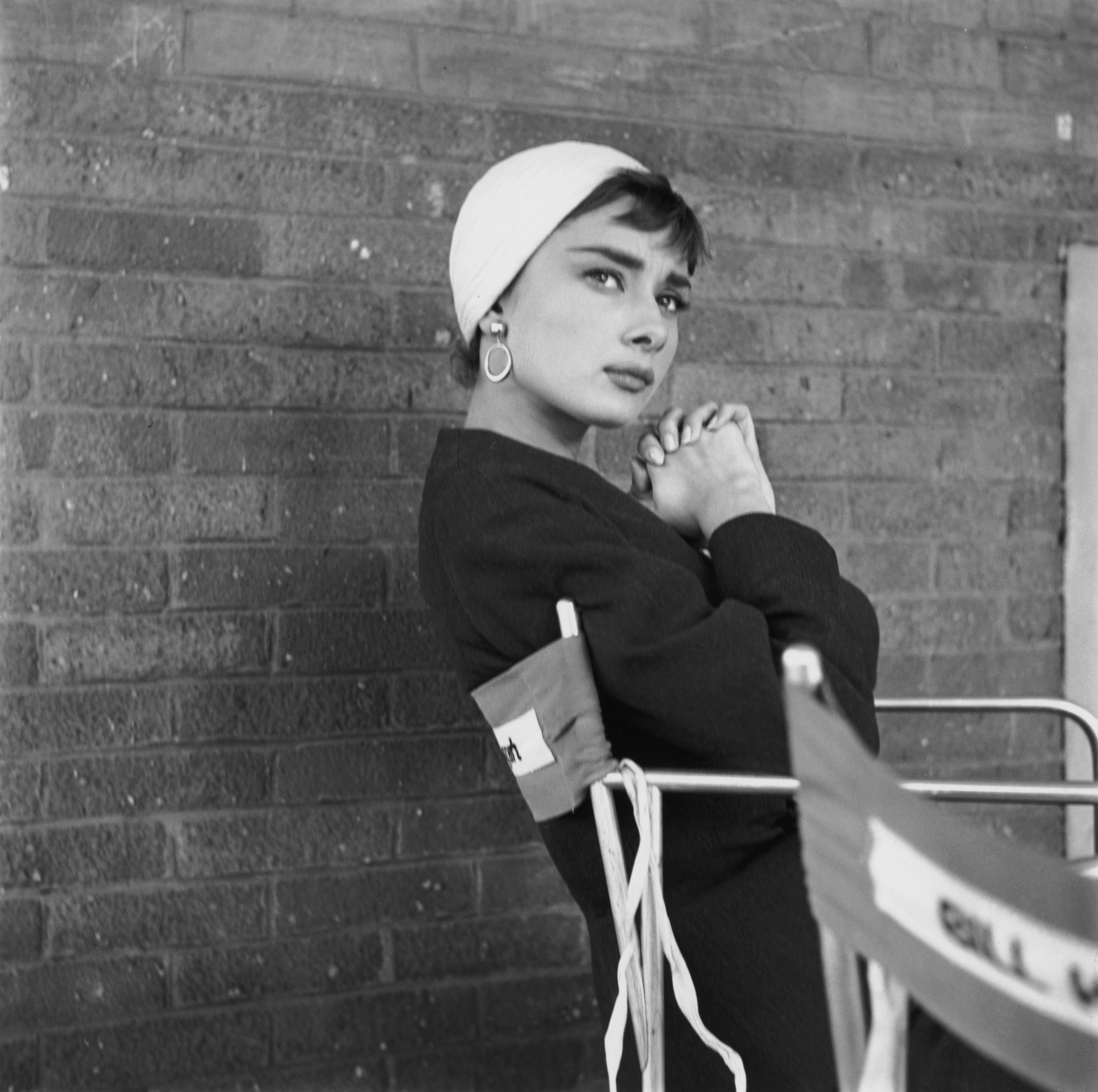 Person in chic vintage style, wearing a hat and earrings, posing against a brick wall, exuding elegance