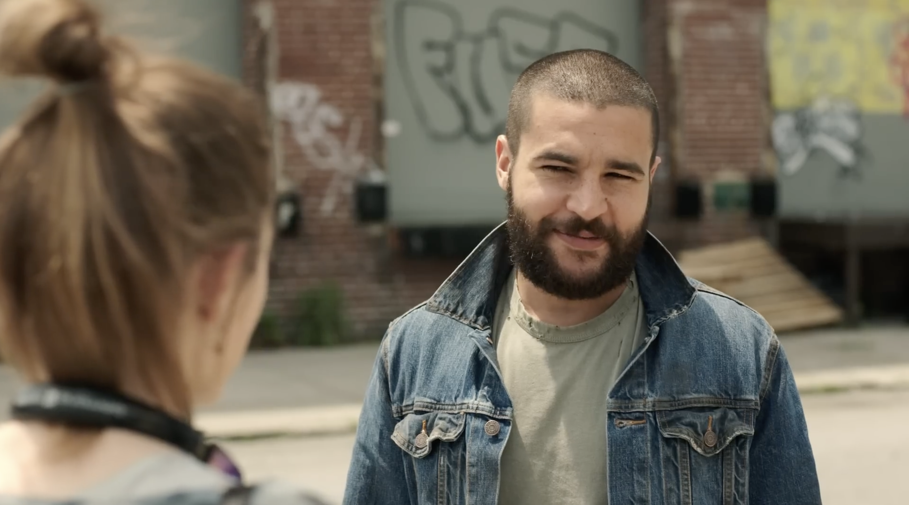 Person with short hair and beard wearing a denim jacket, standing outdoors, talking to another person with hair in a bun. Graffiti visible in background