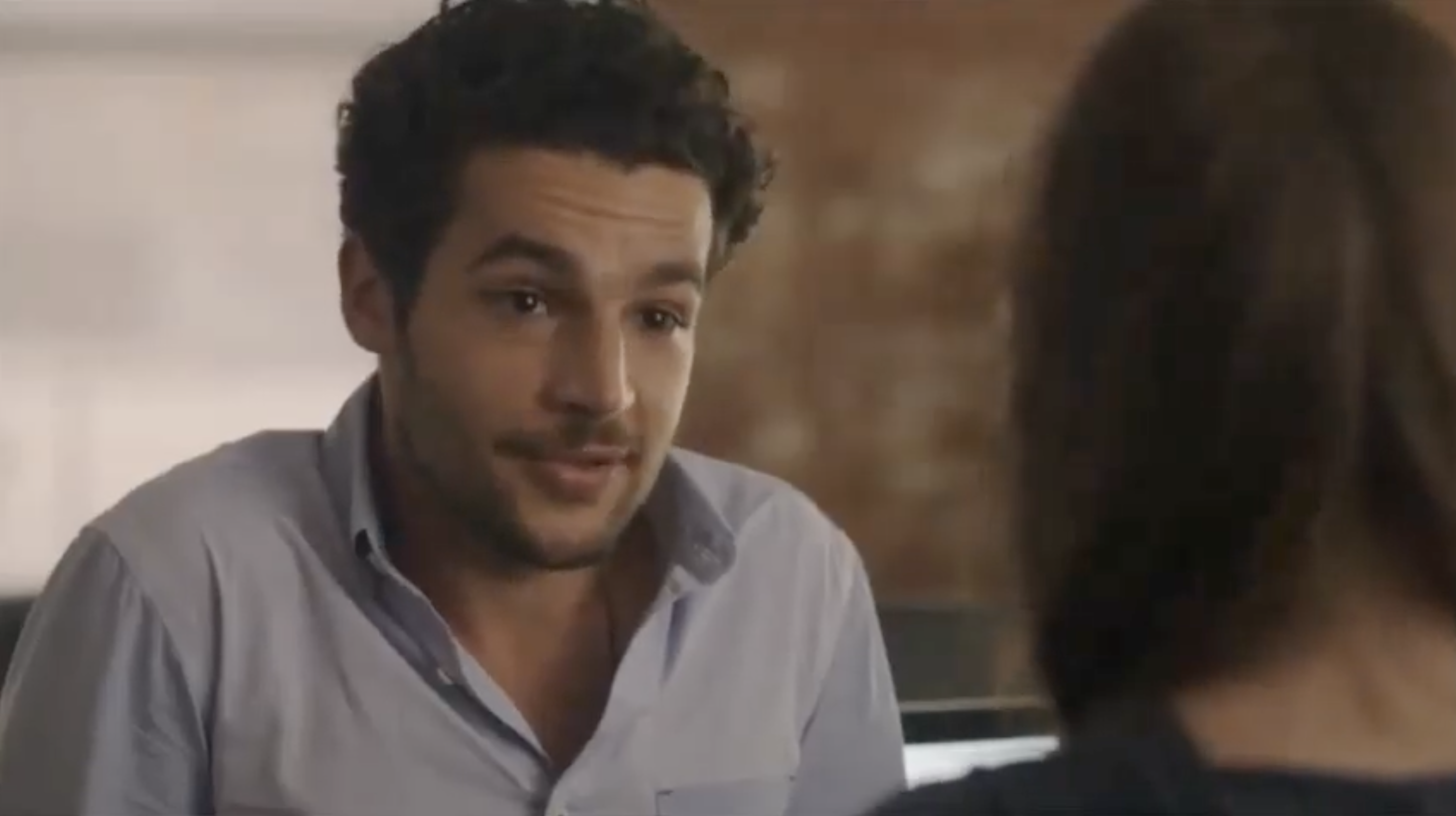 A man in a casual shirt speaks to a woman who is turned away, indoor setting