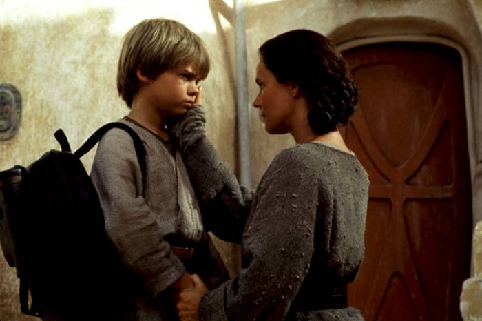 A woman gently touches a boy's face, both in plain textured clothing, outside a rustic building. The moment conveys care and emotion