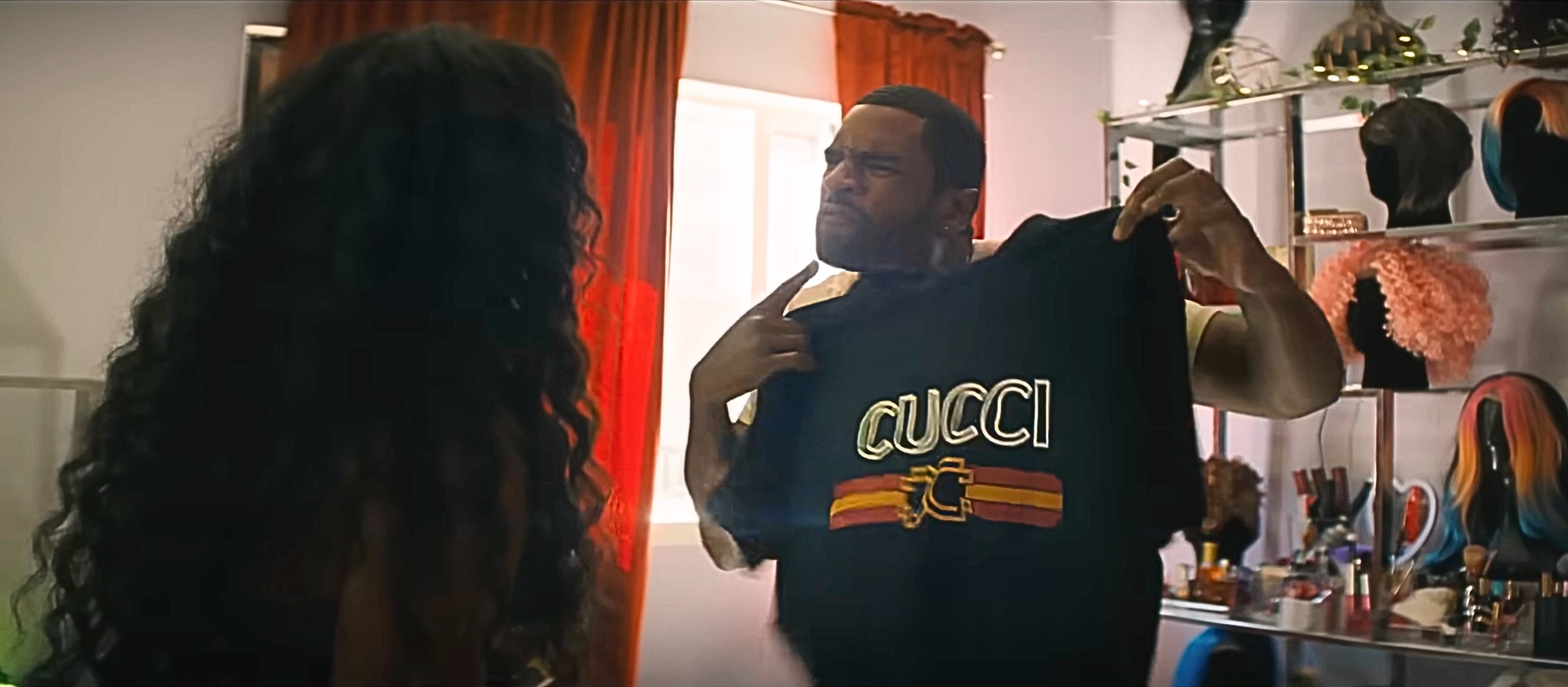 A man humorously holds up a Gucci shirt, pointing at it. A woman with long hair stands nearby, partially turned away