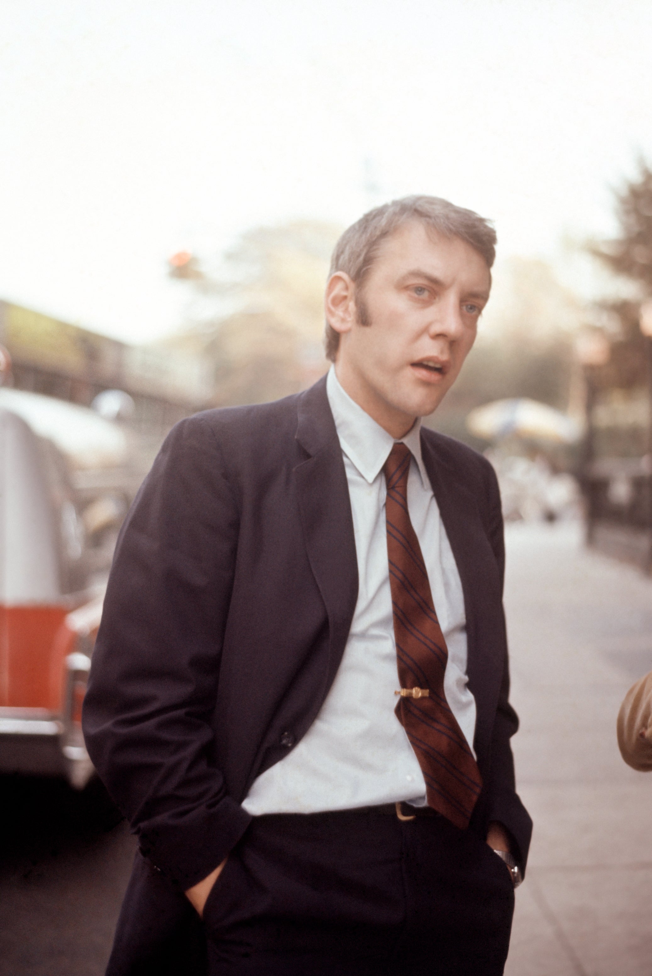 Person in a suit with a tie, standing outdoors on a sidewalk, hands in pockets, looking engaged or thoughtful