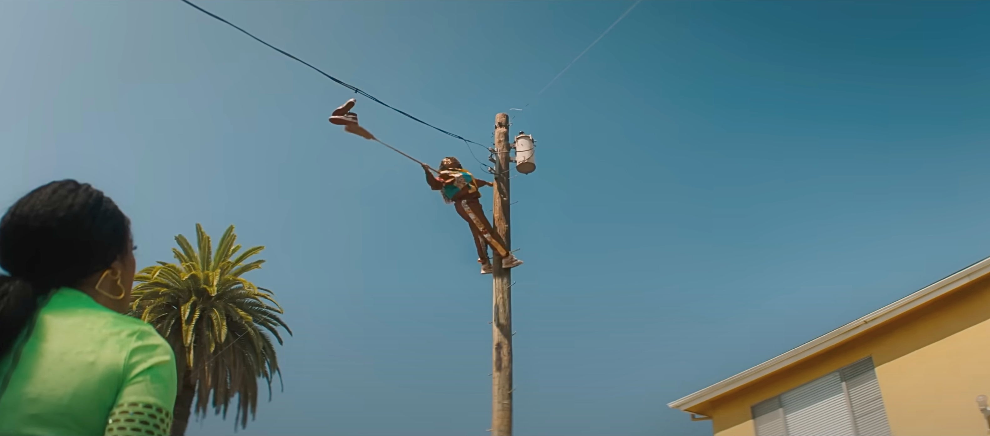 Person climbing a utility pole holding shoes, watched by another person. Palm tree and yellow building in the background