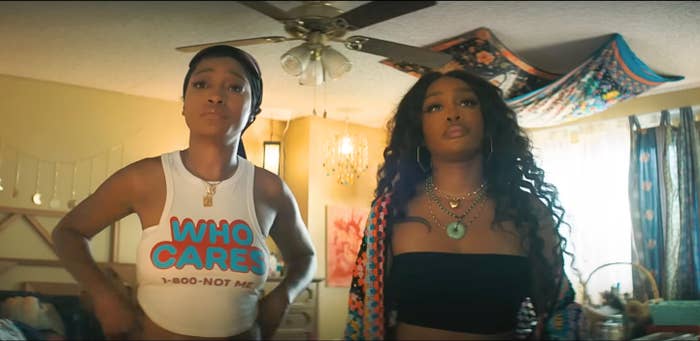 Two individuals in a casual setting. One wears a tank top with "WHO CARES" text; the other wears a black tube top. Both have serious expressions