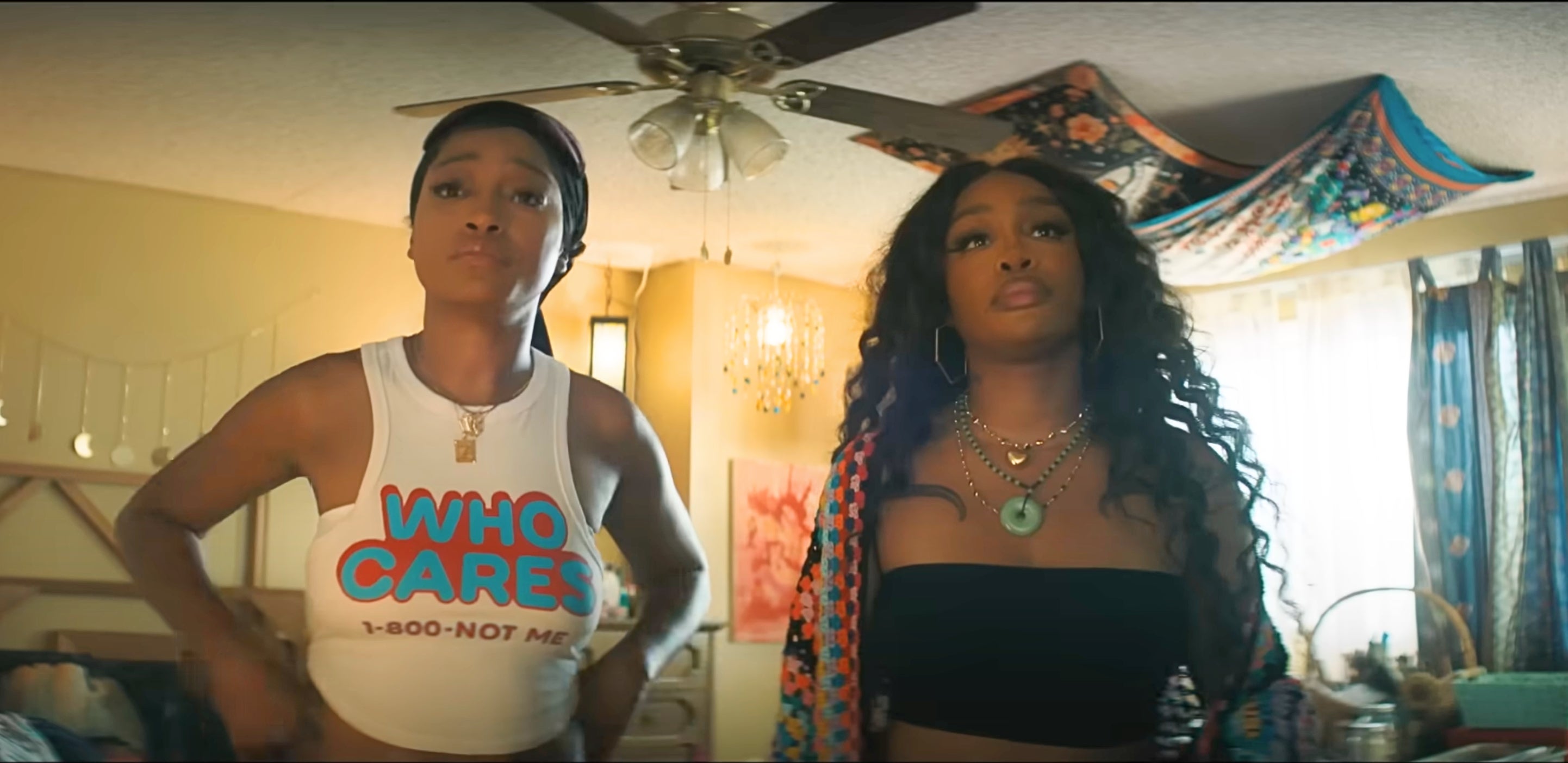 Two women in a room, one wearing a "Who Cares? 1-800-NOT-ME" tank top, the other in a black top with a floral shawl draped over her shoulders