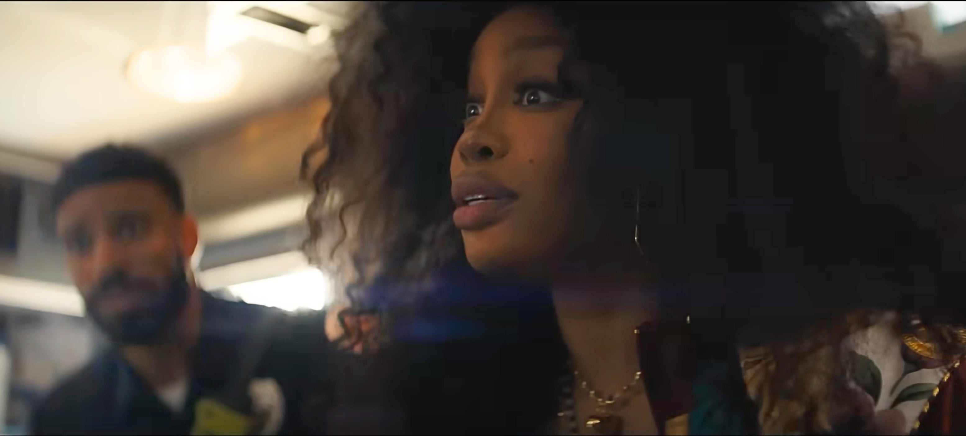 A person with voluminous, curly hair looks surprised in a candid indoor setting with another person in the background