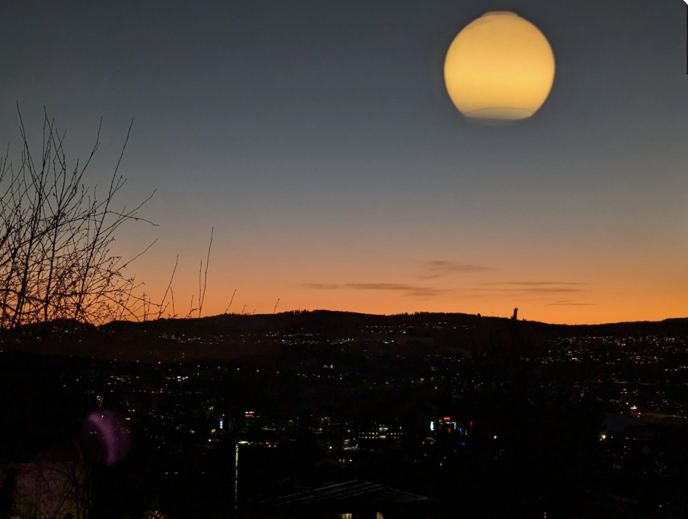 Night cityscape with a sunset horizon, distant hills, and bright lights; a round light fixture is visible in the top right corner of the image