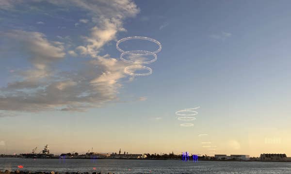 Circular light patterns hover over a body of water, with ships and buildings on the horizon under a partly cloudy sky