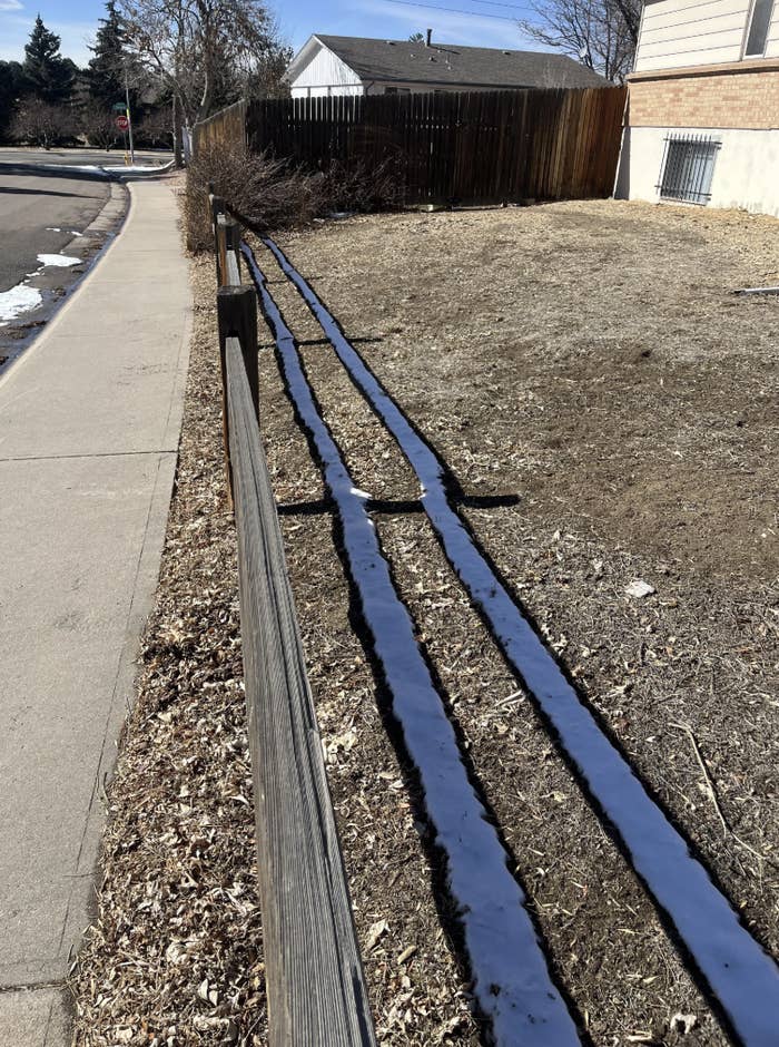 Two lines of snow melt beside a wooden fence on a dry ground near a sidewalk; sparse snow patches visible around