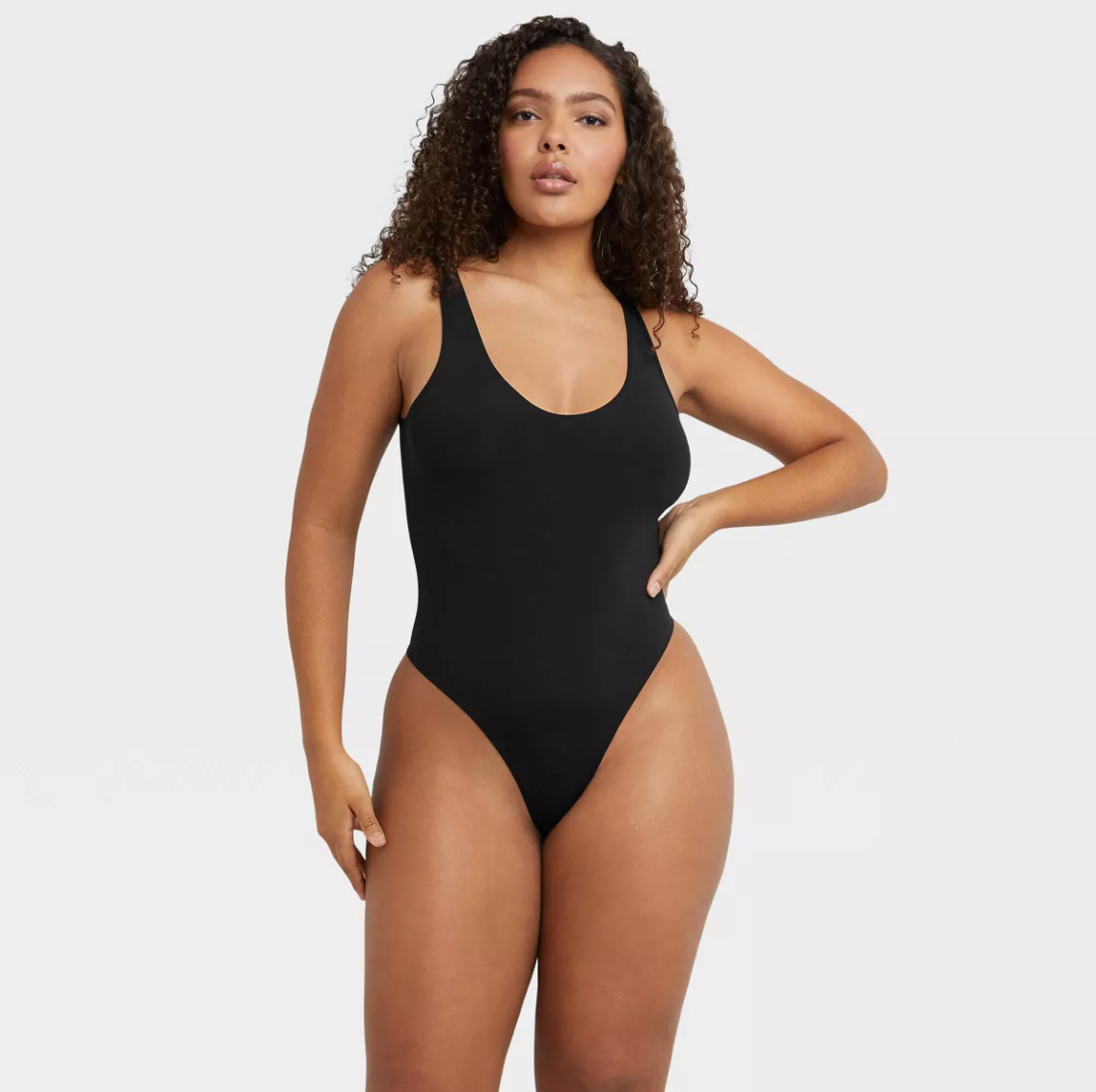 Person in a sleek, form-fitting black one-piece swimsuit, standing confidently with one hand on hip against a neutral background