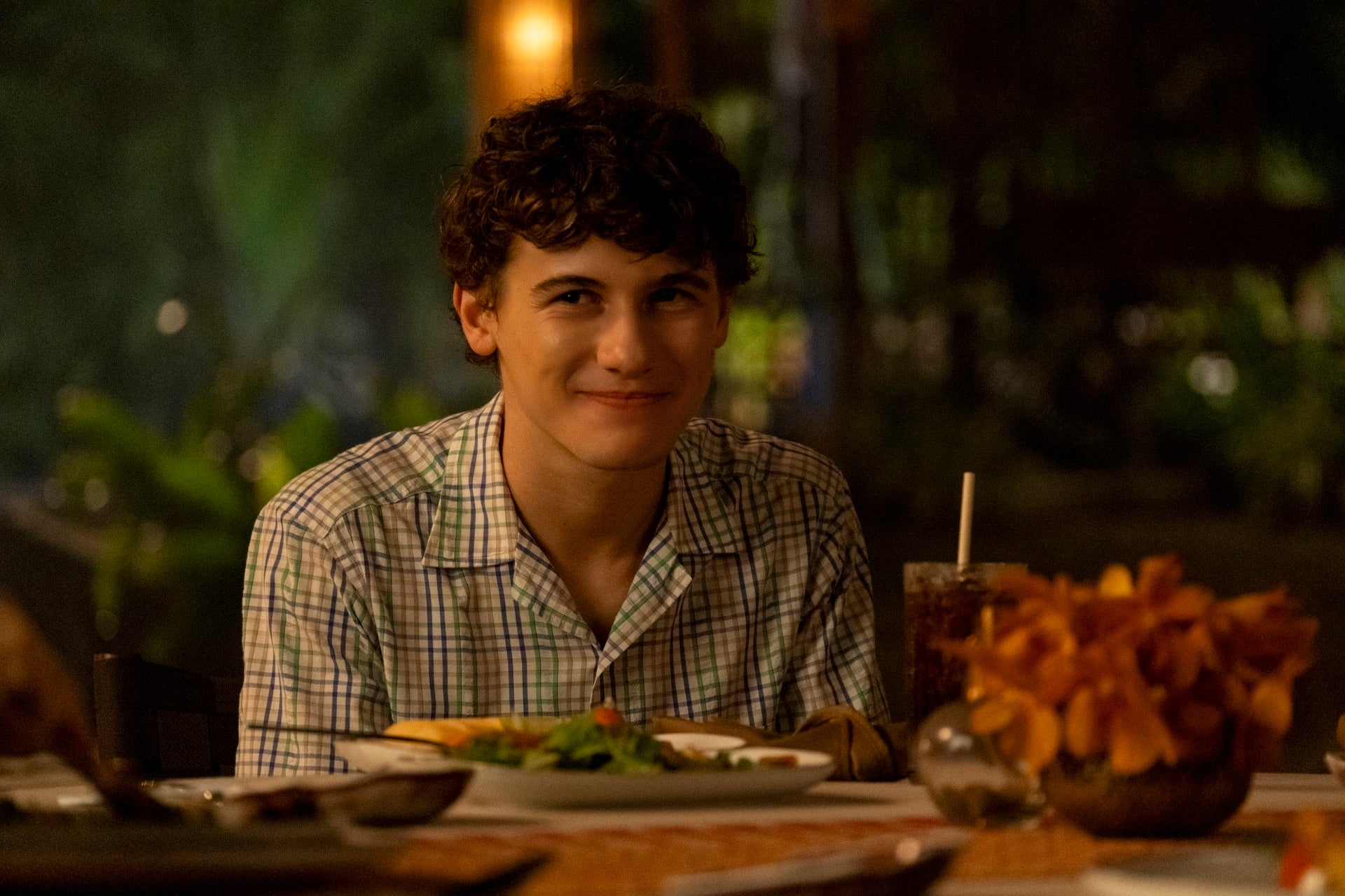 A person smiles at a dinner table with a plaid shirt on, surrounded by food and drink, in a cozy setting