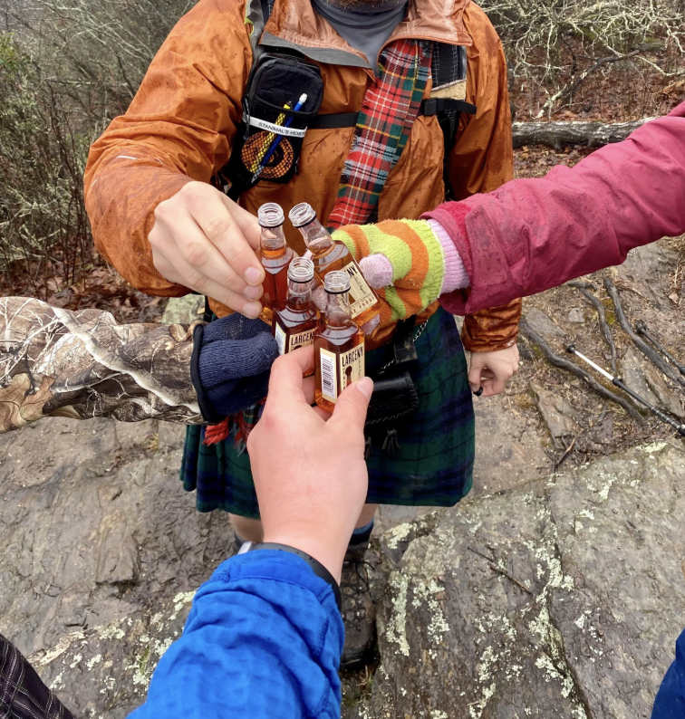 People in outdoor attire toast with small whiskey bottles on a rocky surface, wearing layered clothing and gloves