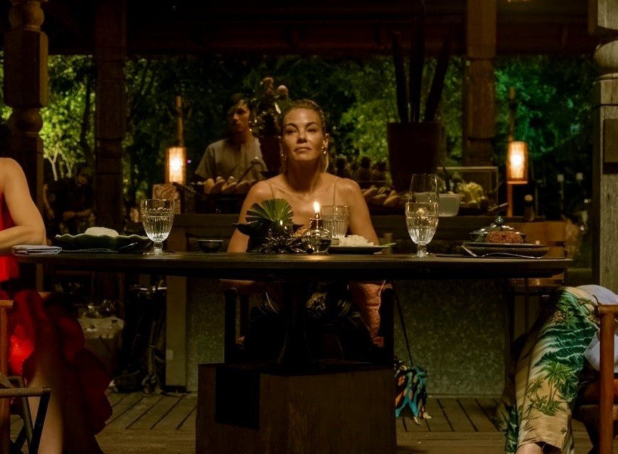 Three people in stylish evening attire are seated at a restaurant table, reacting with surprise. The setting appears tropical and open-air