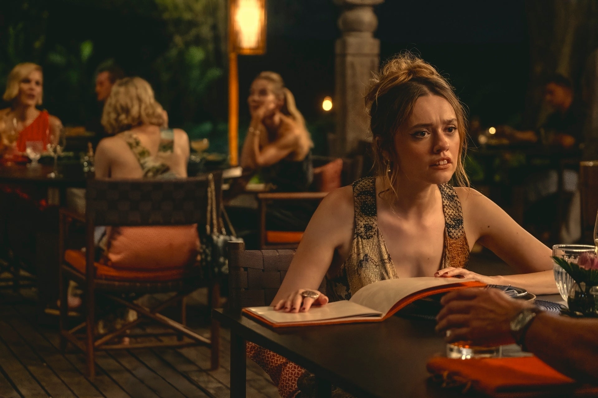 A woman at an outdoor restaurant looks surprised with an open menu in front of her. Other diners are seen in the background