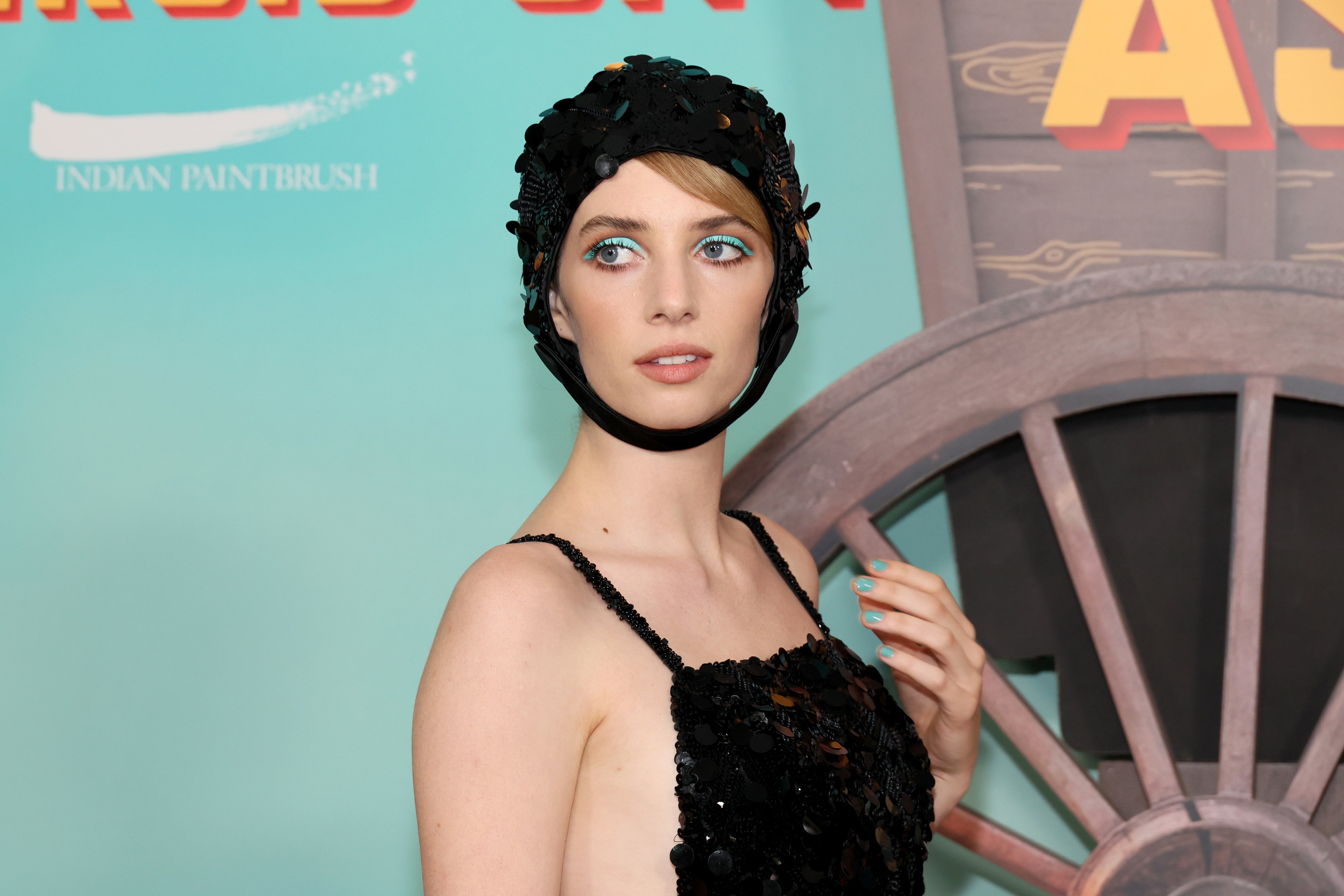 Maya Hawke poses on the red carpet in a black textured dress and headpiece, next to a wagon wheel backdrop