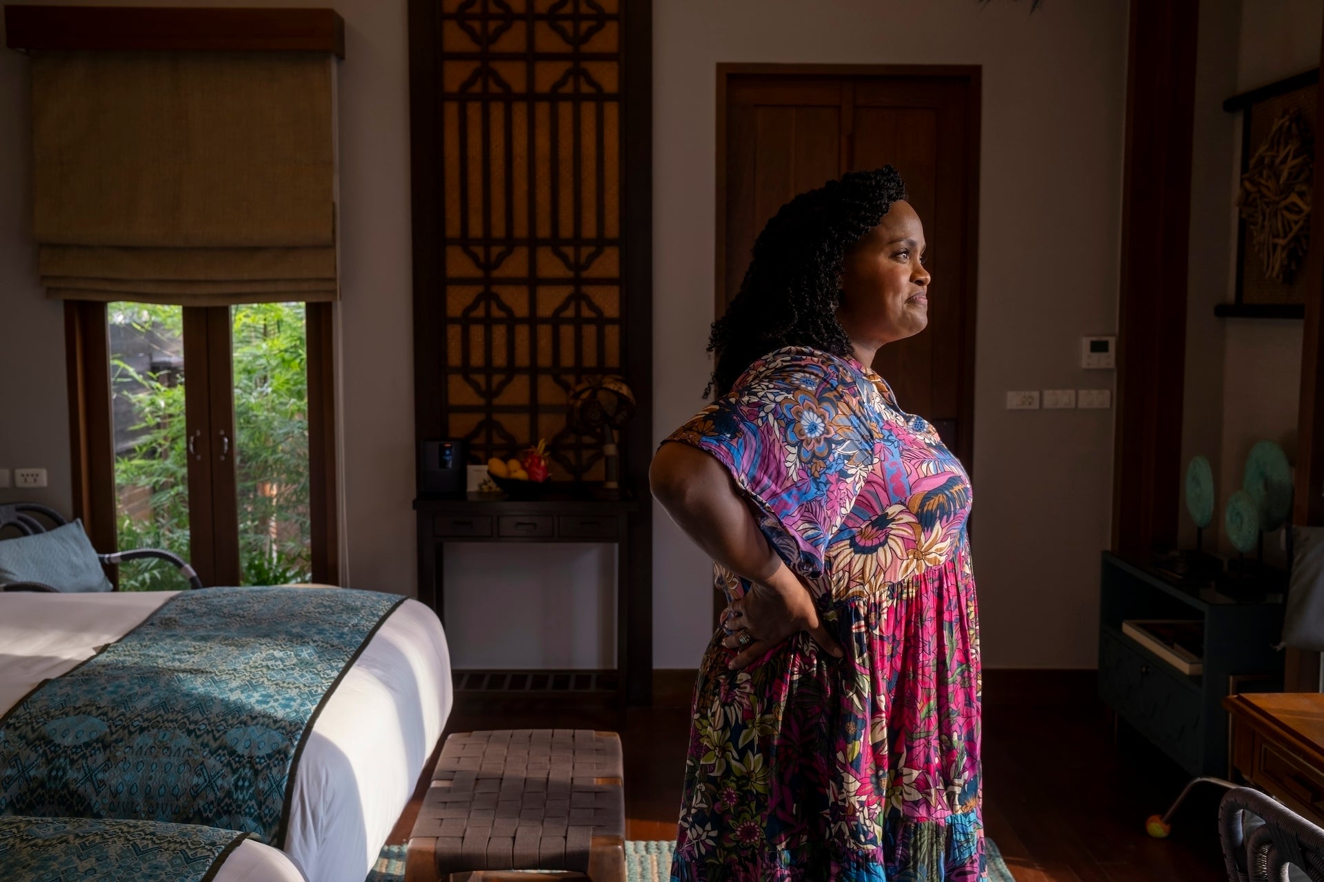 Person stands in a thoughtfully decorated room, wearing a vibrant, floral dress, looking contemplatively out a window